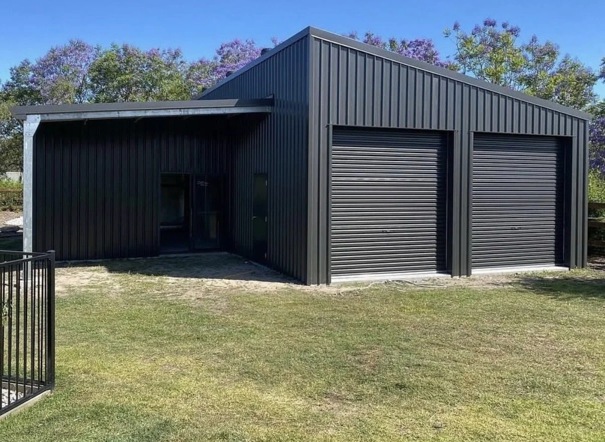 Black metal garage with two roll-up doors, located on a grassy yard, surrounded by trees with purple blossoms.
