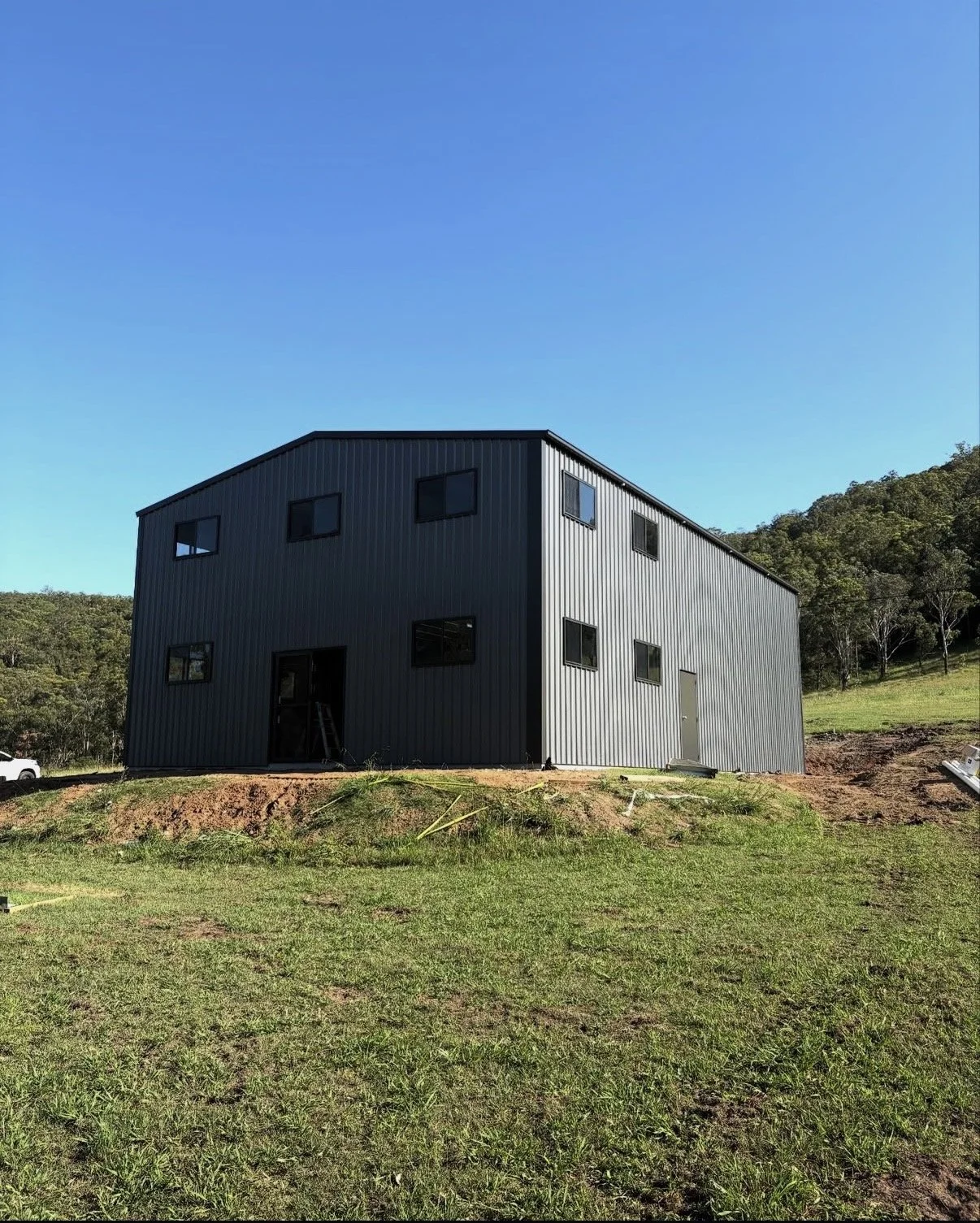 Newly built grey metal house on a grassy hill with trees in the background and a clear blue sky.