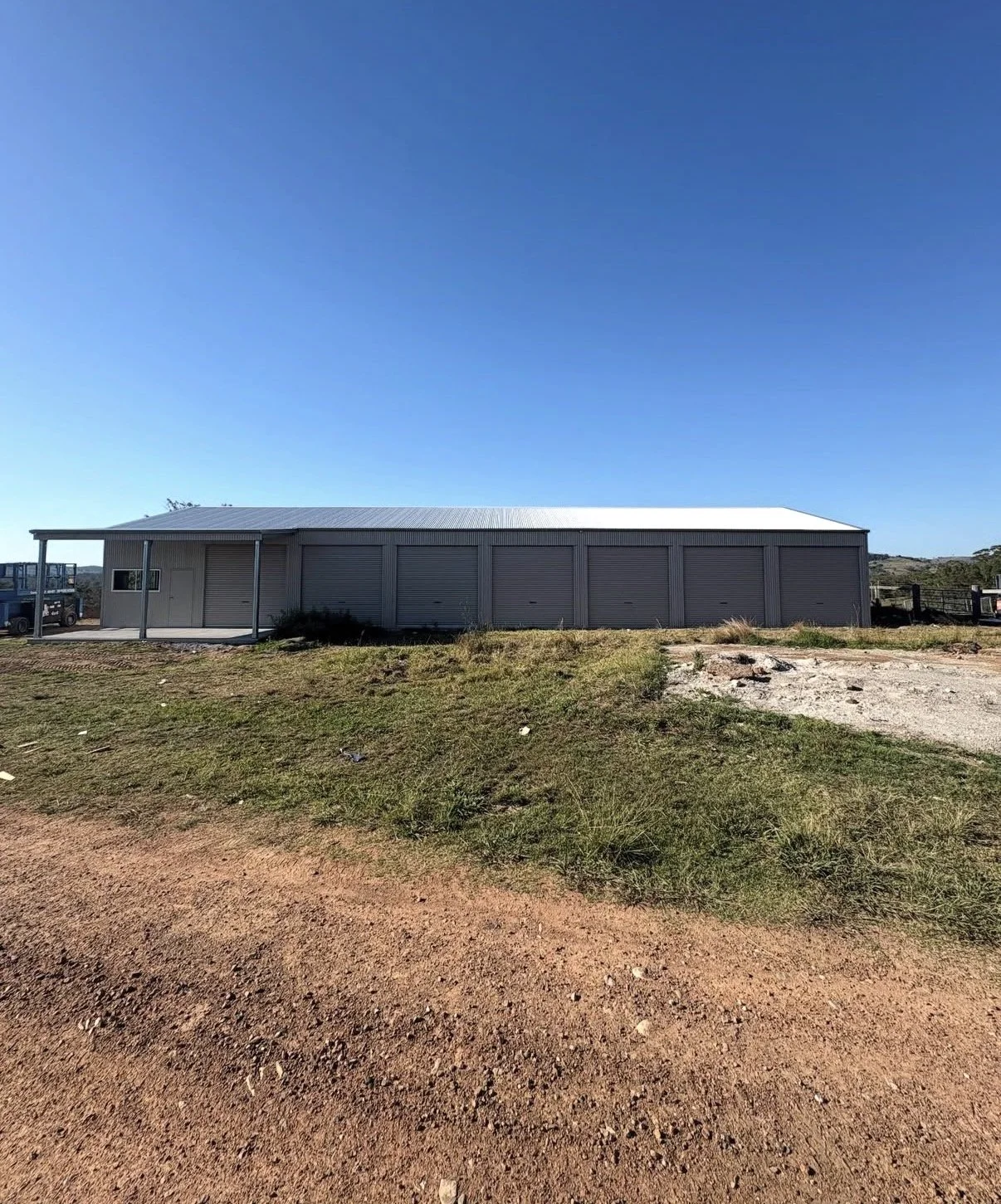 A large, metal industrial building with a white roof and multiple garage doors, located in a rural area with dirt and grass foreground and clear blue sky.