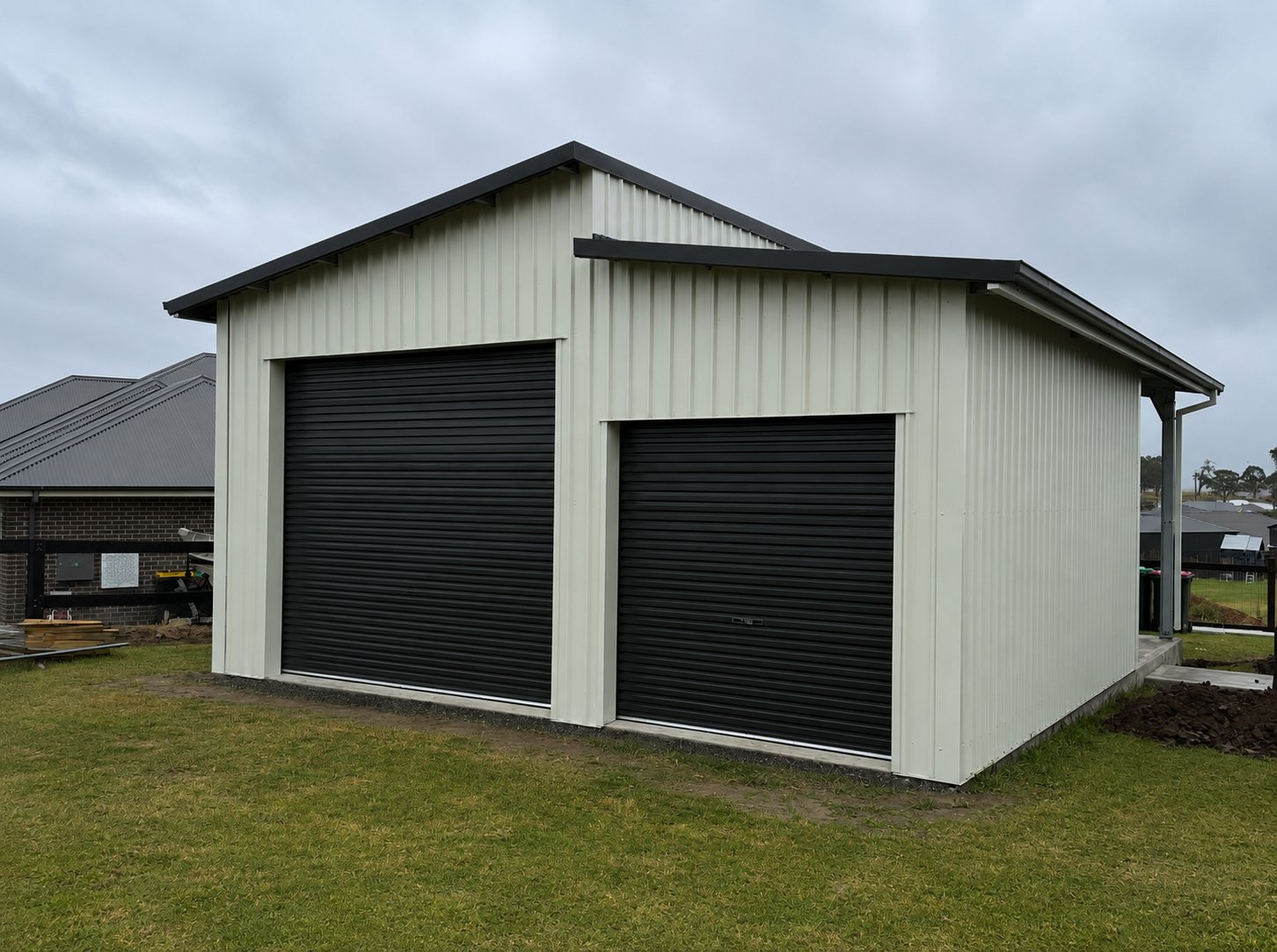 A white metal garage with two black roll-up doors situated on a grassy yard under a cloudy sky.