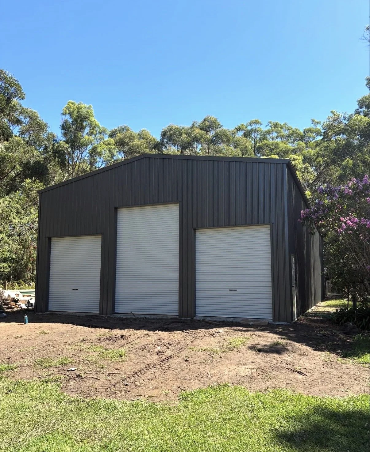 A black metal garage with three white roll-up doors, situated outdoors with trees and a clear blue sky in the background.