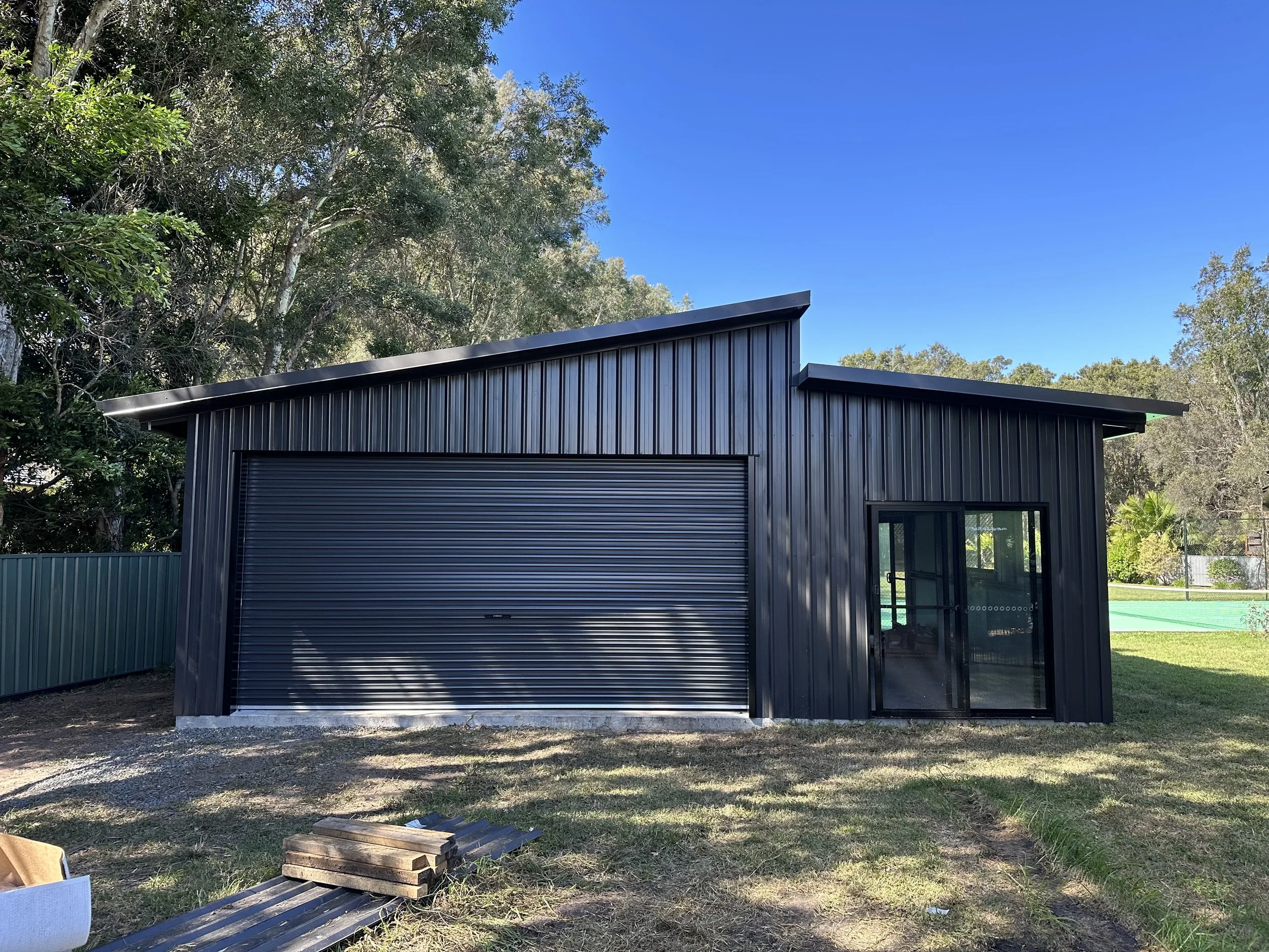 Exterior view of a modern metal building with a closed roller door, set against a background of trees and a clear blue sky.