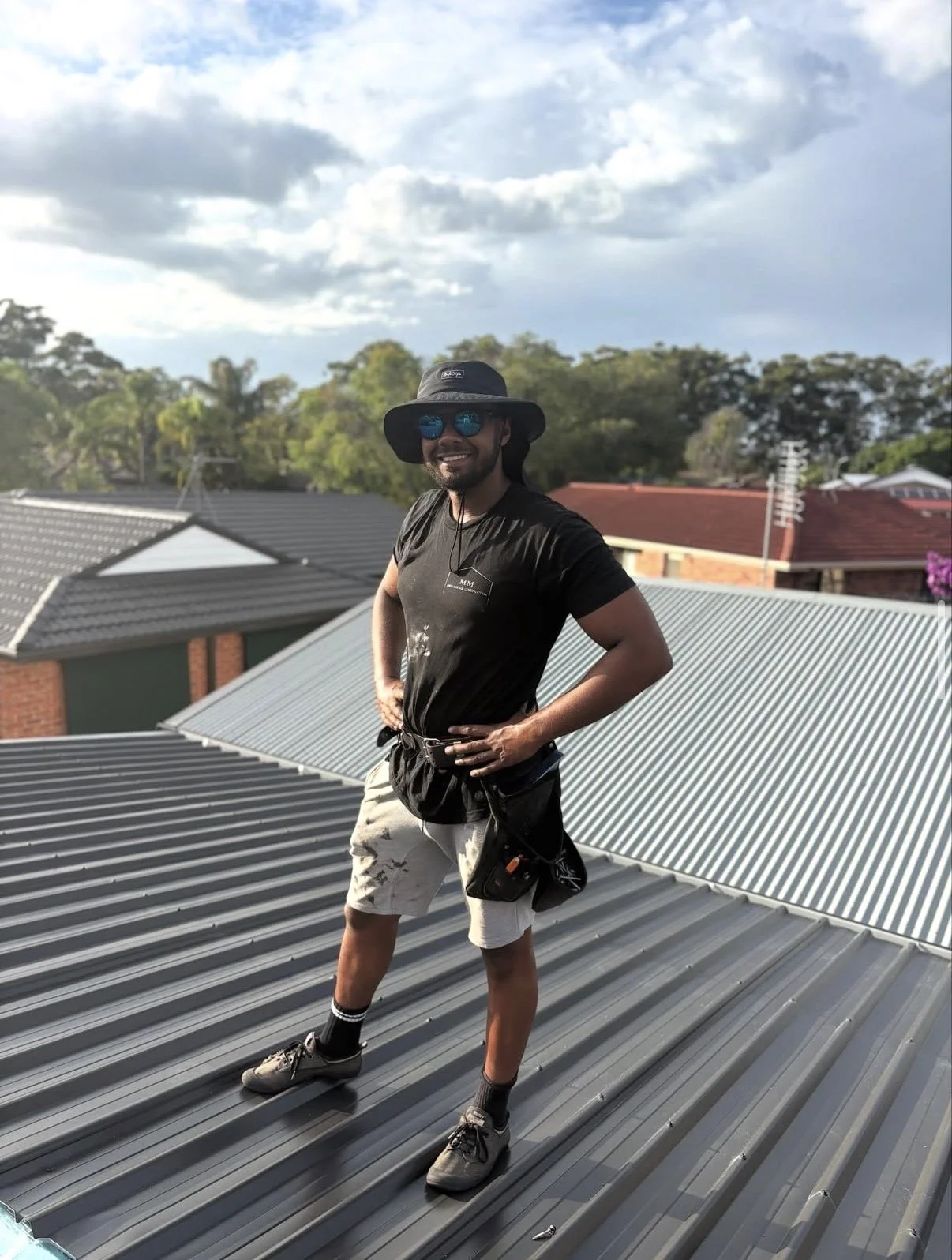 A man standing on a metal roof with his hands on his hips, wearing sunglasses, a wide-brimmed hat, a black t-shirt, beige shorts, and hiking shoes, smiling at the camera with trees and a partly cloudy sky in the background.