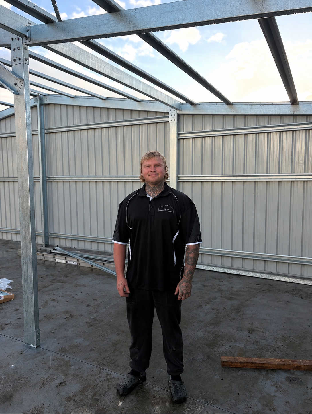 A young man with tattoos on his neck and arms standing inside a metal framework structure on a construction site, wearing a black sports shirt and black pants.