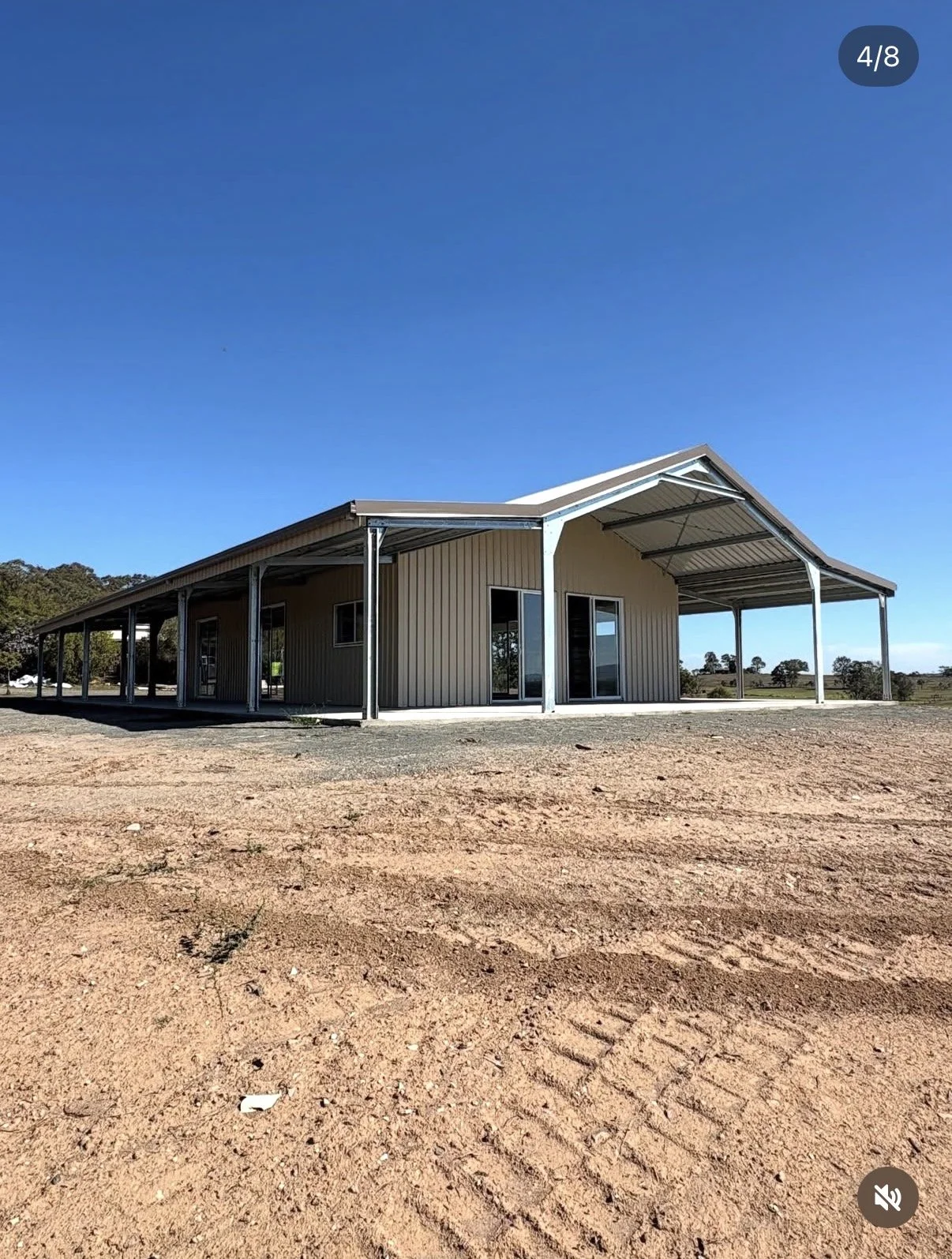 A large metal building with a covered porch, set on a dirt lot under a clear blue sky. The building has vertical metal siding, multiple windows and glass doors.