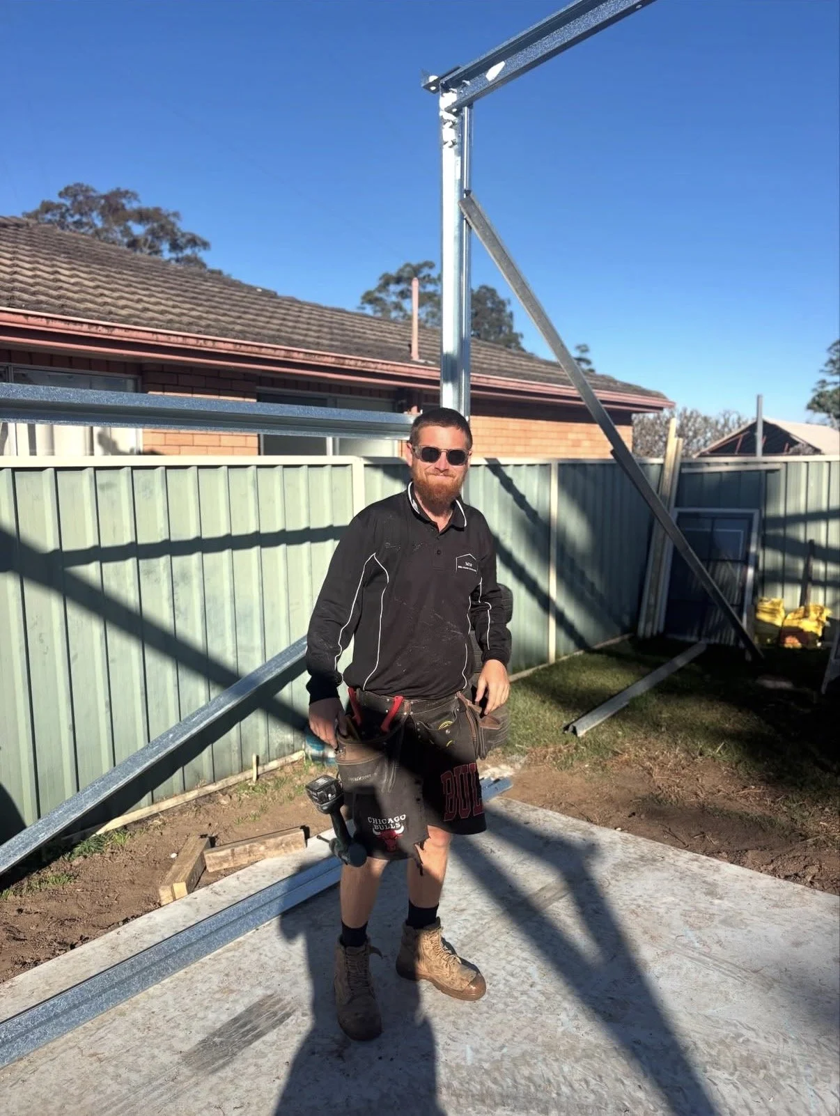 A man with sunglasses, a black long-sleeve shirt, shorts, and work boots standing on a construction site with metal framework being assembled.