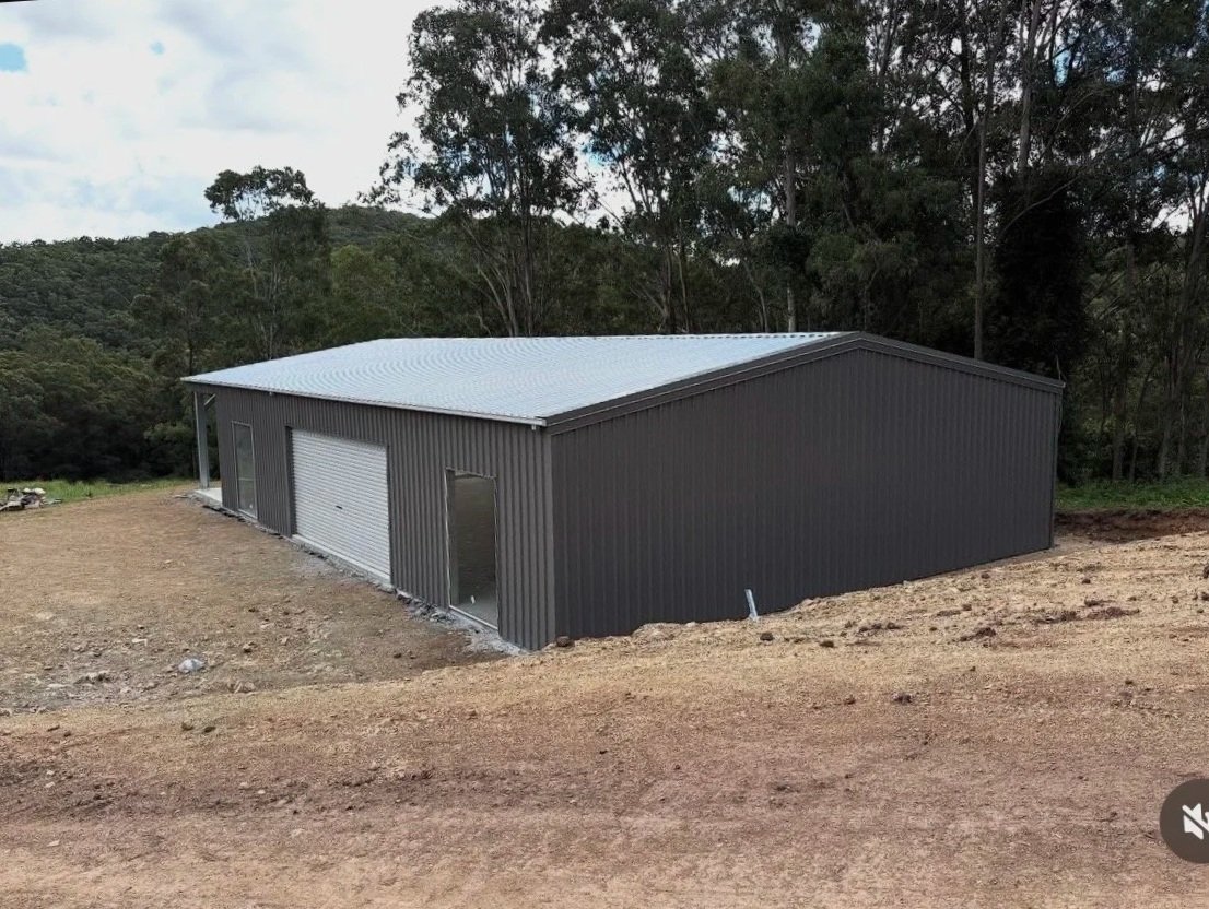 A metal building with a silver roof and dark gray walls situated on a dirt lot near a wooded area.