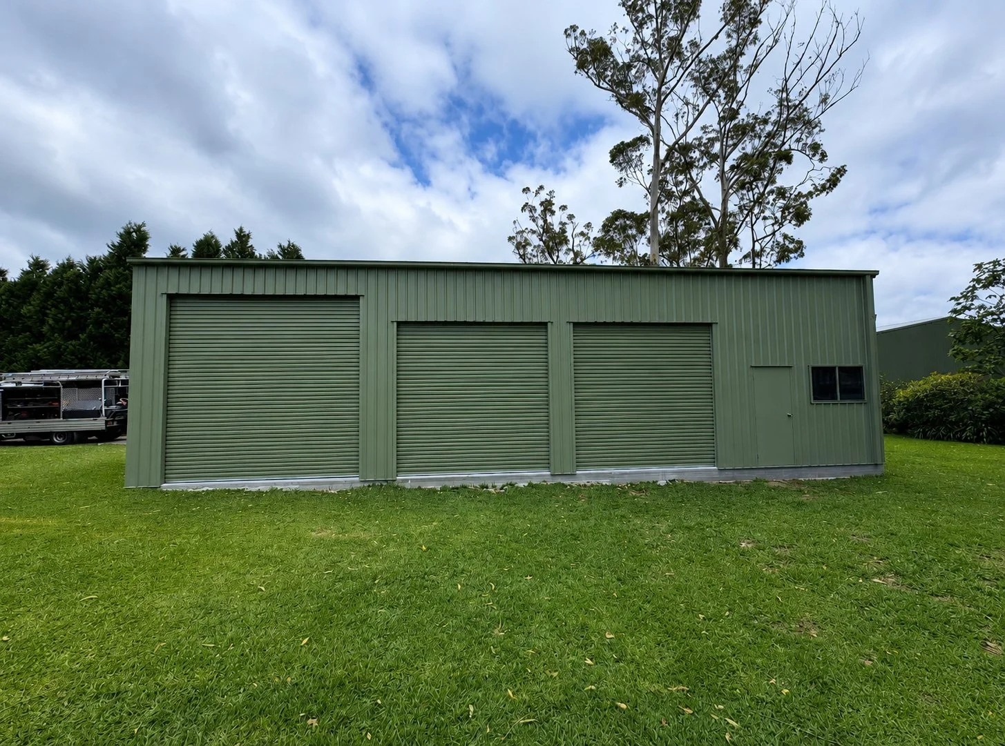 A green metal storage shed with three rolling doors and a small window on the right side, situated on a grassy lawn with trees and a partly cloudy sky in the background.