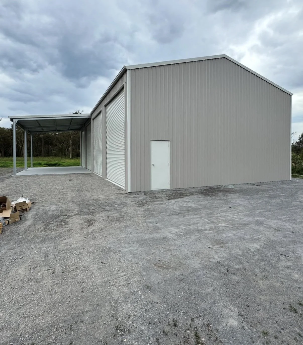 A large metal storage building with a small door and a partially open garage door on a gravel lot, surrounded by trees under a cloudy sky.