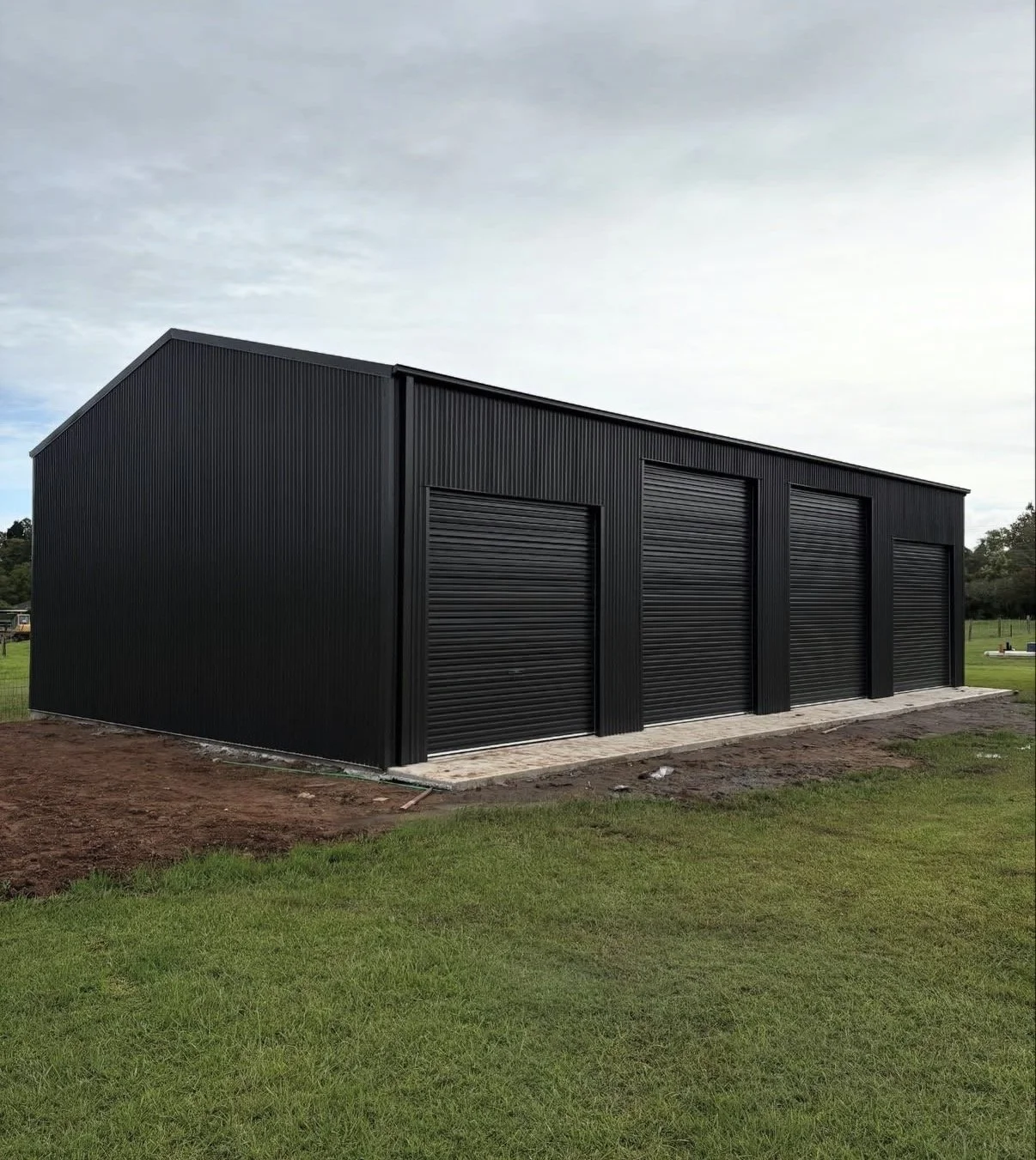 Black metal storage building with three roll-up garage doors, situated on a grassy field with a cloudy sky overhead.