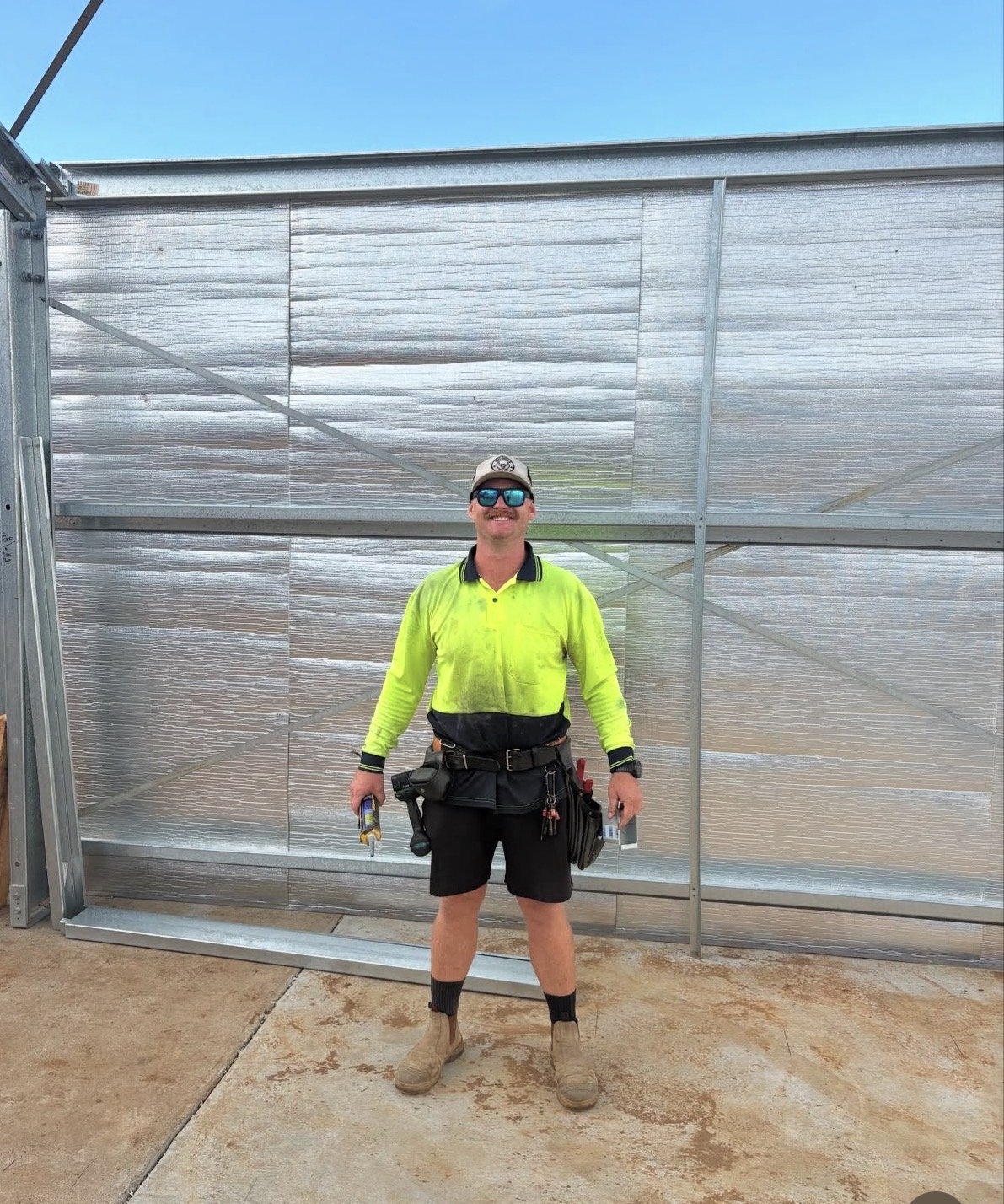 A smiling construction worker in a high-visibility yellow shirt, black shorts, and safety gear, standing in front of a metallic fence on a construction site.