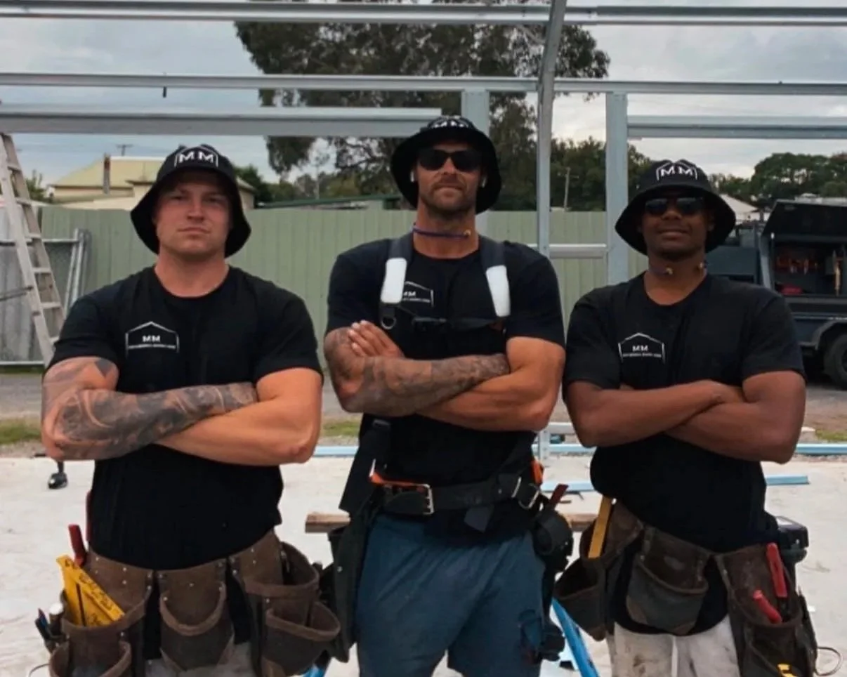 Three construction workers standing with arms crossed in front of a metal framework, wearing black shirts, tool belts, and hats with 'M M' logo.