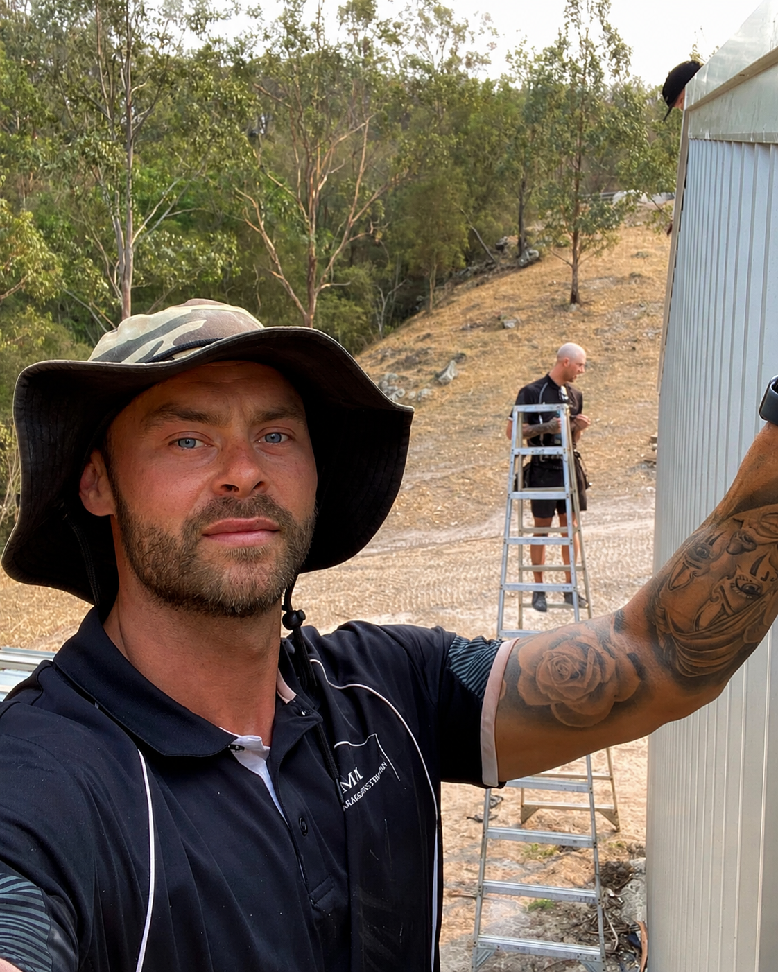 A man in a wide-brimmed hat and black shirt taking a selfie outdoors, with two other men working on a building wall in the background near a ladder and a wooded area.