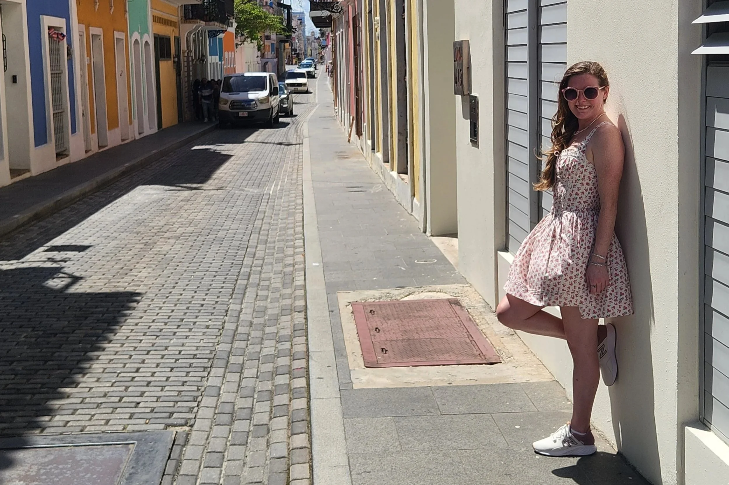 A young woman with long hair wearing pink sunglasses, a floral dress, and sneakers, leaning against a white wall on a colorful street with parked cars and buildings in the background.
