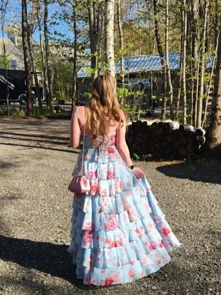 A woman with long, wavy red hair wearing a strapless, tiered, floral dress in pink, blue, and white. She carries a small purple purse with a gold strap and stands outdoors on a sunny day, with trees and a stack of firewood visible in the background.