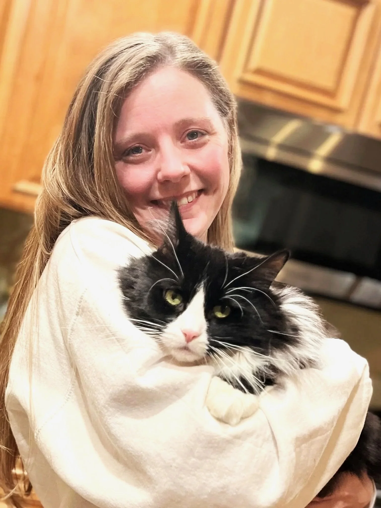 A woman with long blonde hair smiling, holding a black and white cat with yellow eyes, in a kitchen with wooden cabinets in the background.