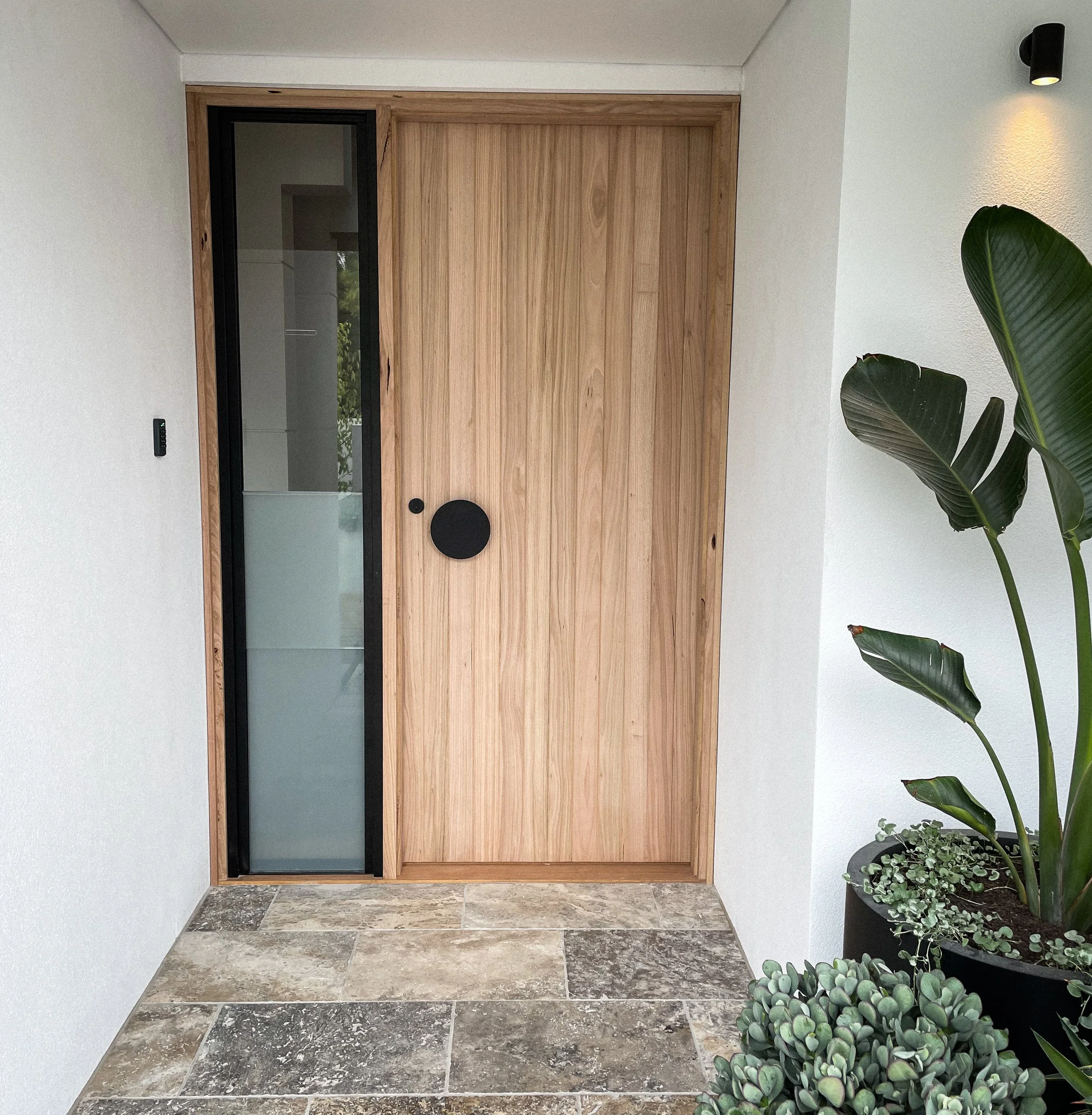 A modern front door with wood paneling and black-framed glass, flanked by white walls, with a small potted plant on the right and tiled stone flooring.