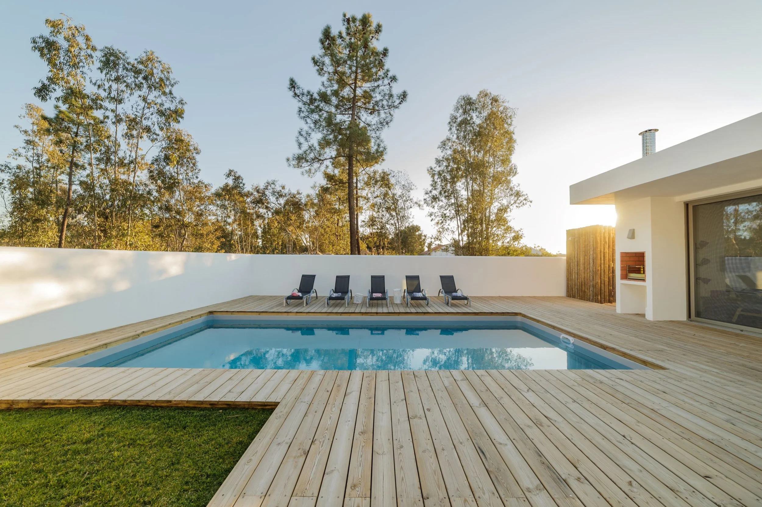 A modern backyard with a swimming pool surrounded by a wooden deck, outdoor lounge chairs, and a white wall. There are green trees and a clear sky in the background during sunset.