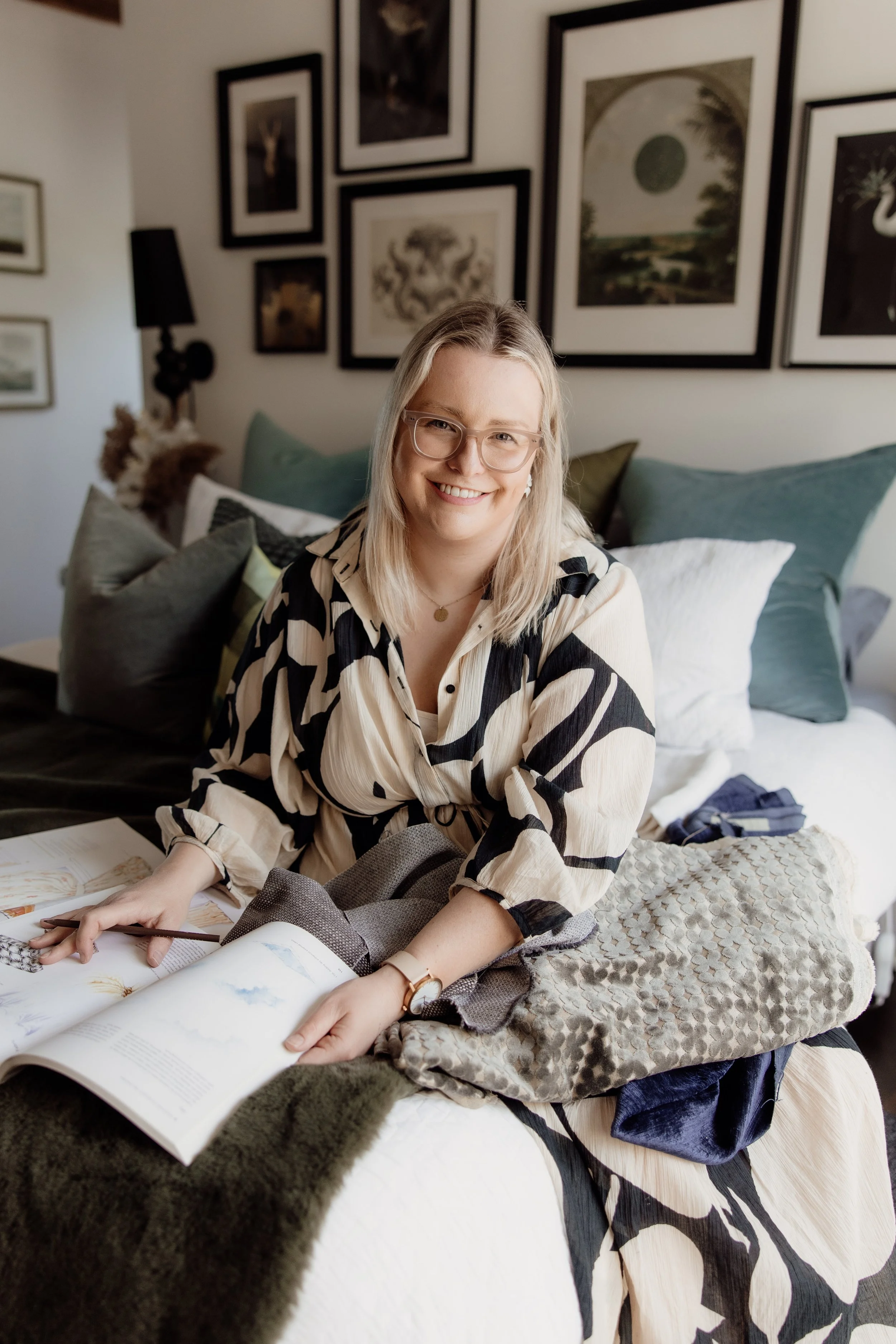 A woman with blonde hair and glasses sitting on a bed, smiling at the camera, surrounded by artwork on the wall and various pillows and textiles on the bed.