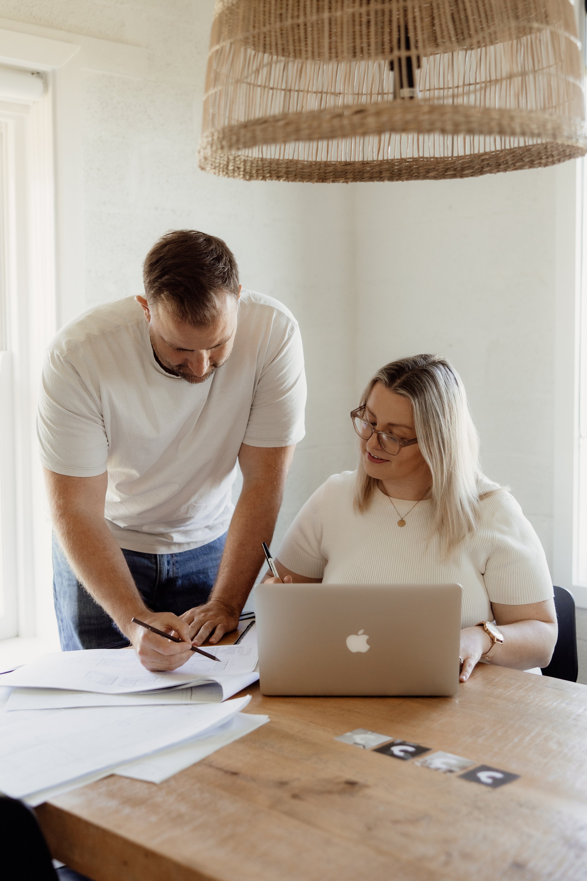 A man and woman working together at a desk, with the man leaning over and pointing at papers, and the woman sitting and using a laptop, in a bright room with a wicker lampshade hanging from the ceiling.
