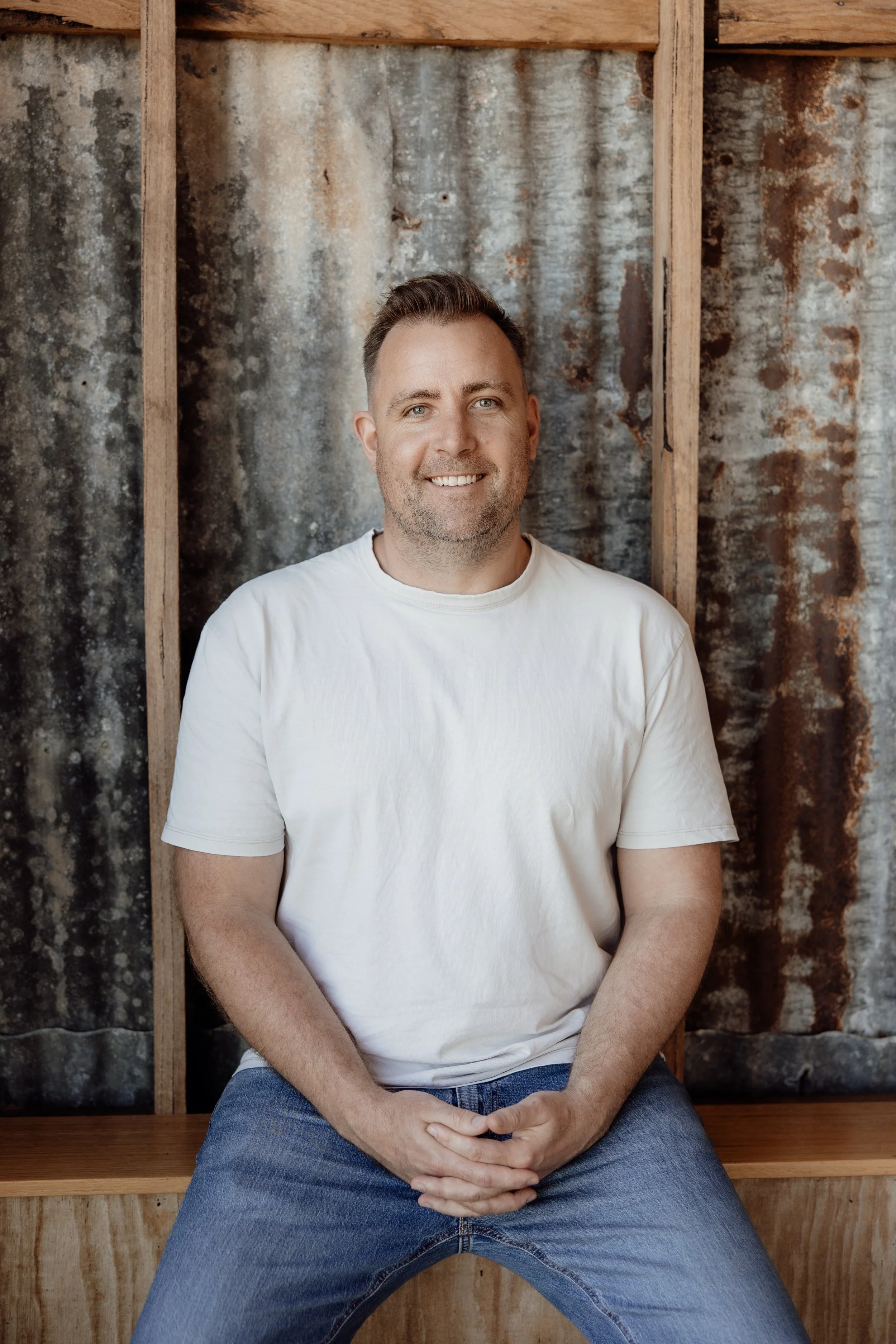 A man sitting on a wooden bench in front of a rustic, weathered metal wall, wearing a white t-shirt and jeans, smiling at the camera.