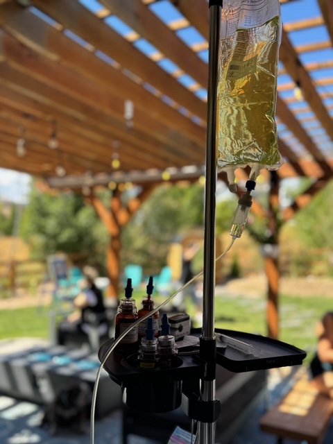 Medical IV drip attached to a stand outdoors with a shaded wooden pergola and picnic table in background.