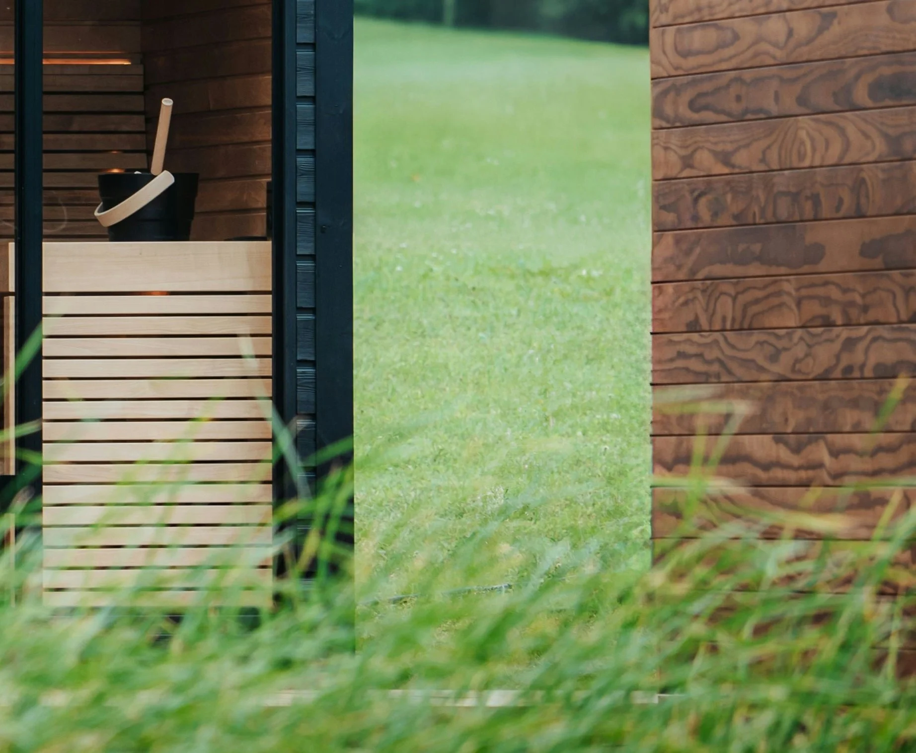 Wooden garden shed with a black bucket and handle visible through an open doorway, surrounded by grass