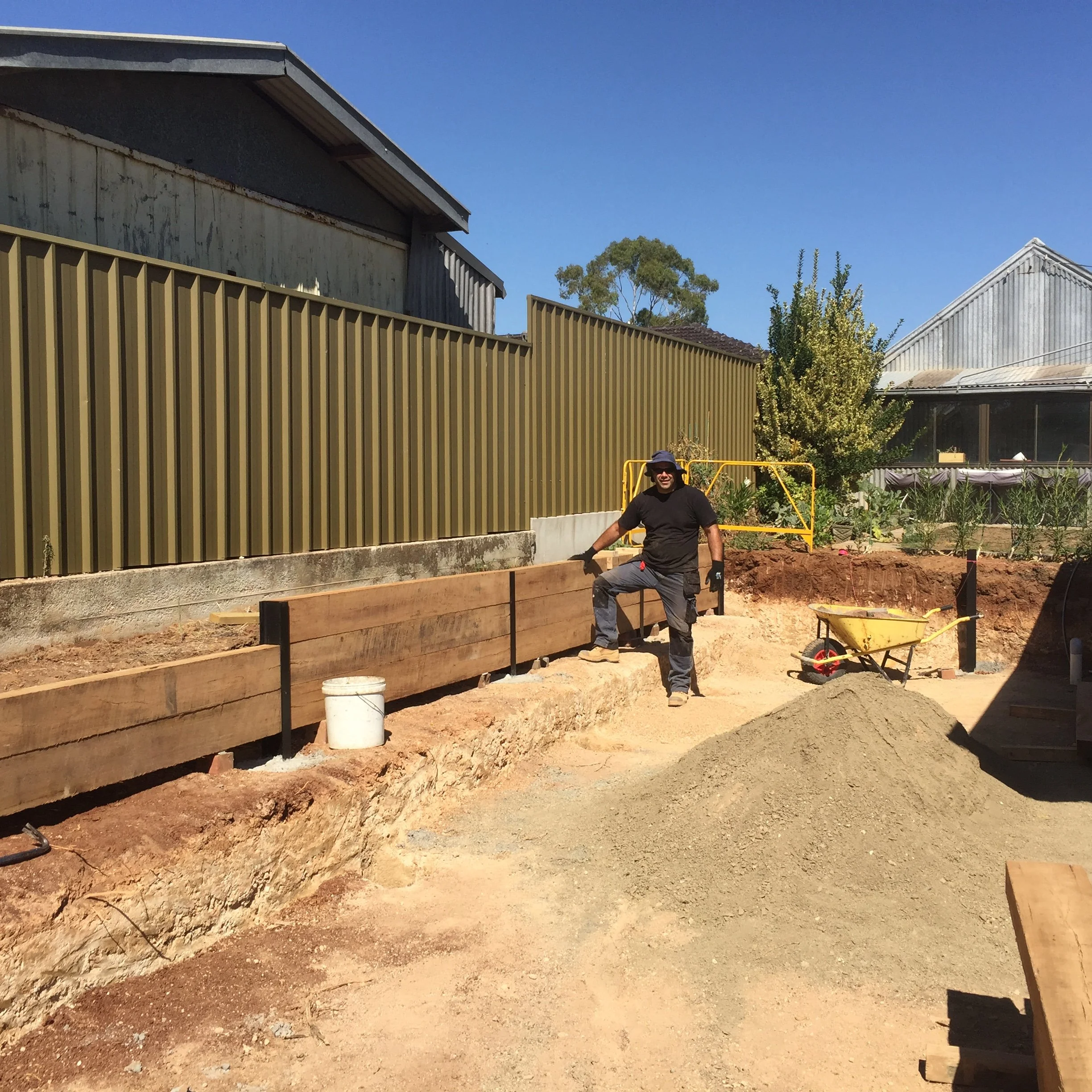 Construction worker building raised garden beds beside a corrugated metal fence, with wheelbarrows and a sunny backyard.