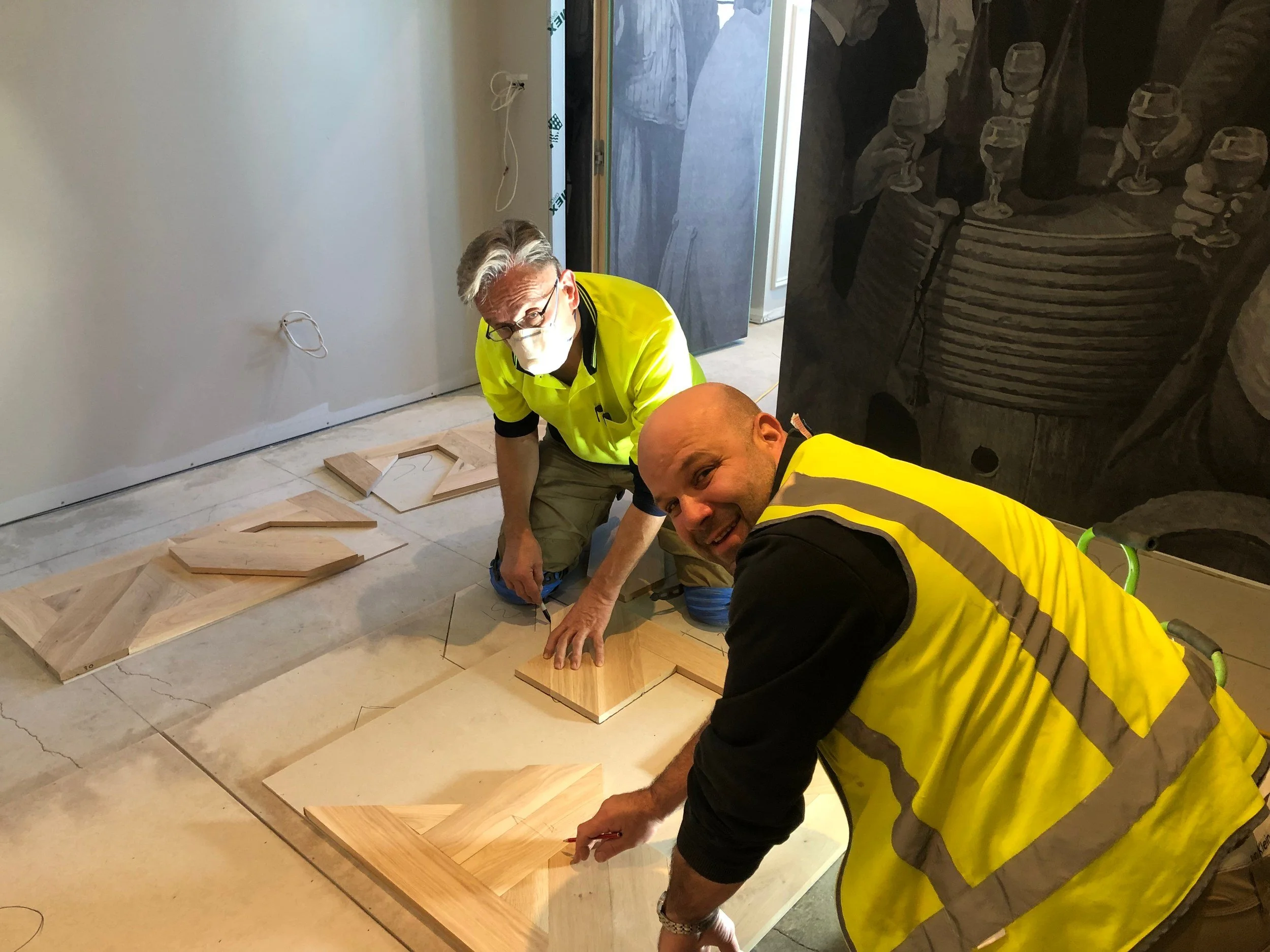 Two construction workers in high-visibility vests installing patterned wood flooring beside a large dinosaur mural.