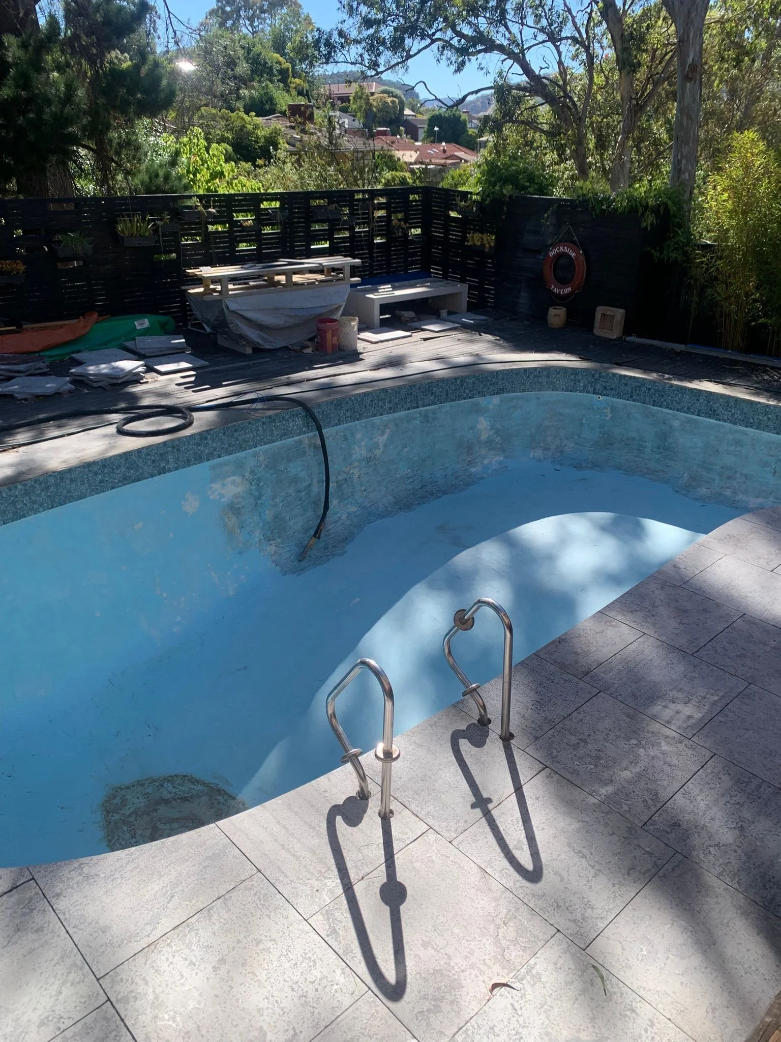 Backyard swimming pool under renovation with exposed pool ladder, patio tiles, outdoor seating, greenery, and suburban homes beyond