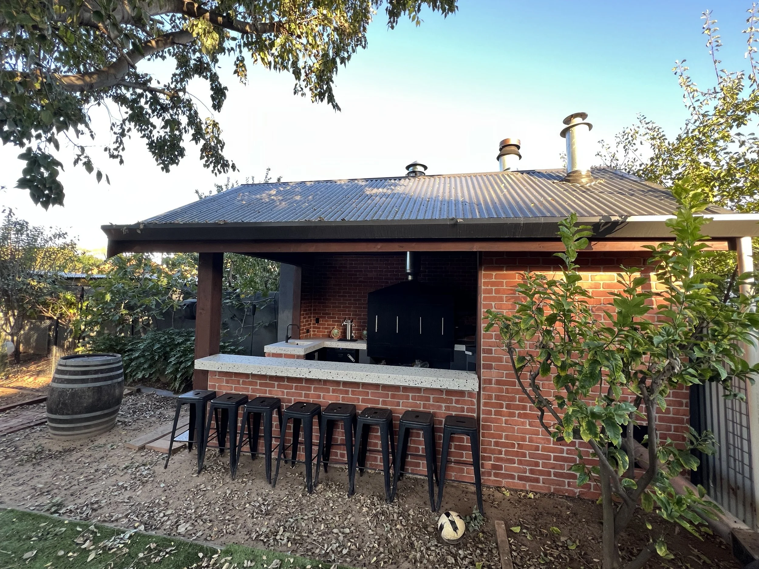 Outdoor brick barbecue kitchen with corrugated metal roof, chimneys, bar counter, stools, and surrounding trees