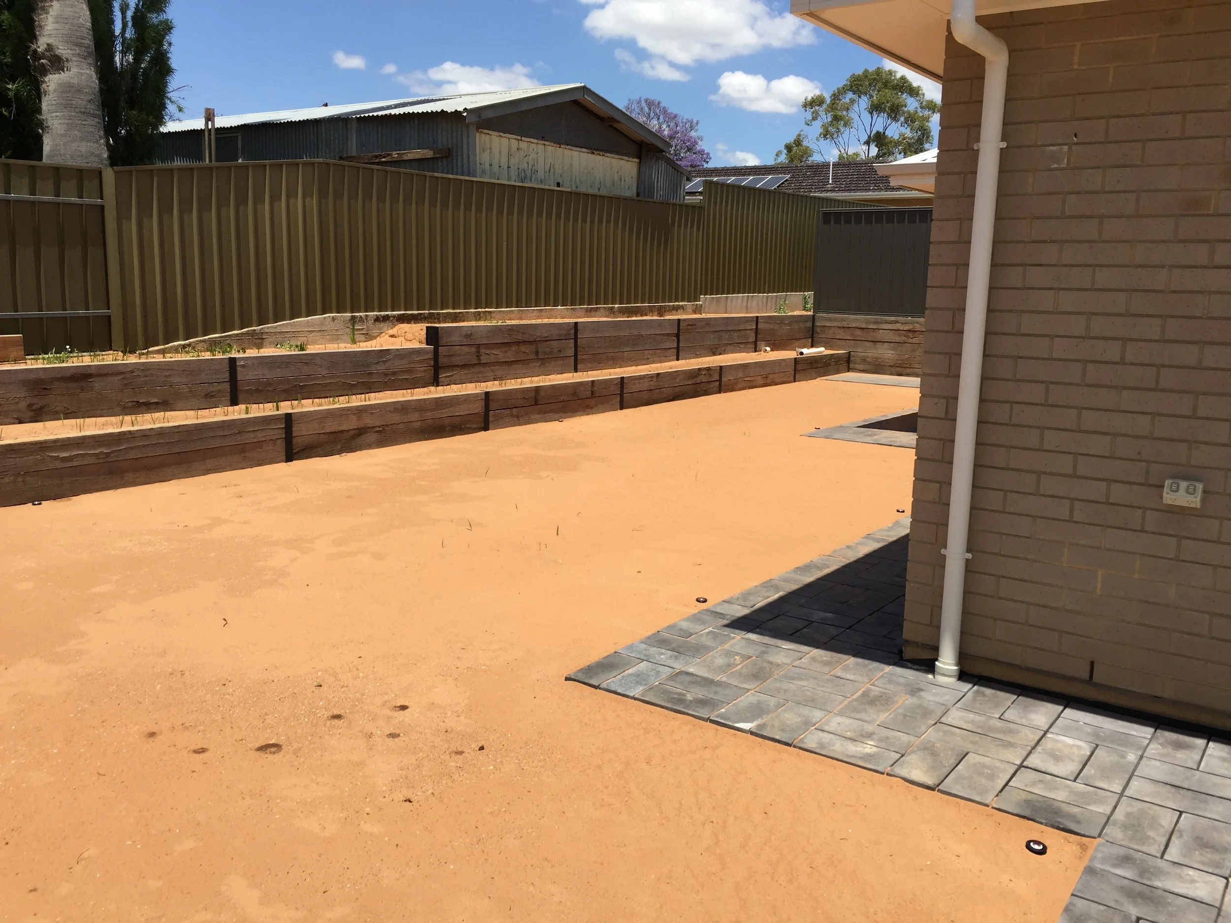 Sunny residential backyard with sandy ground, tiered timber garden beds, corrugated metal fencing, patio pavers, and neighboring sheds