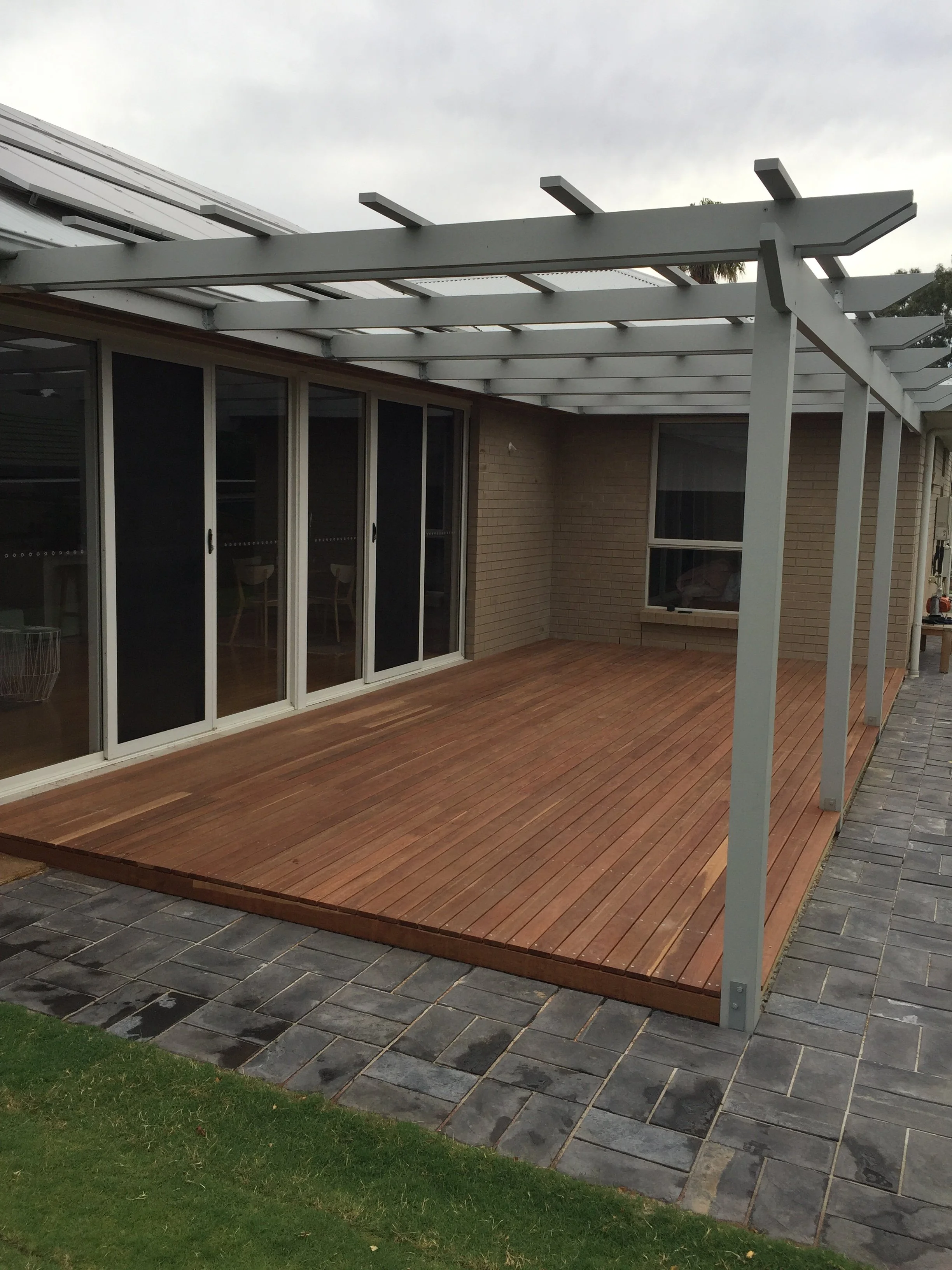 Modern covered patio with a wooden deck, white pergola beams, glass doors, brick walls, and paved stone walkway.