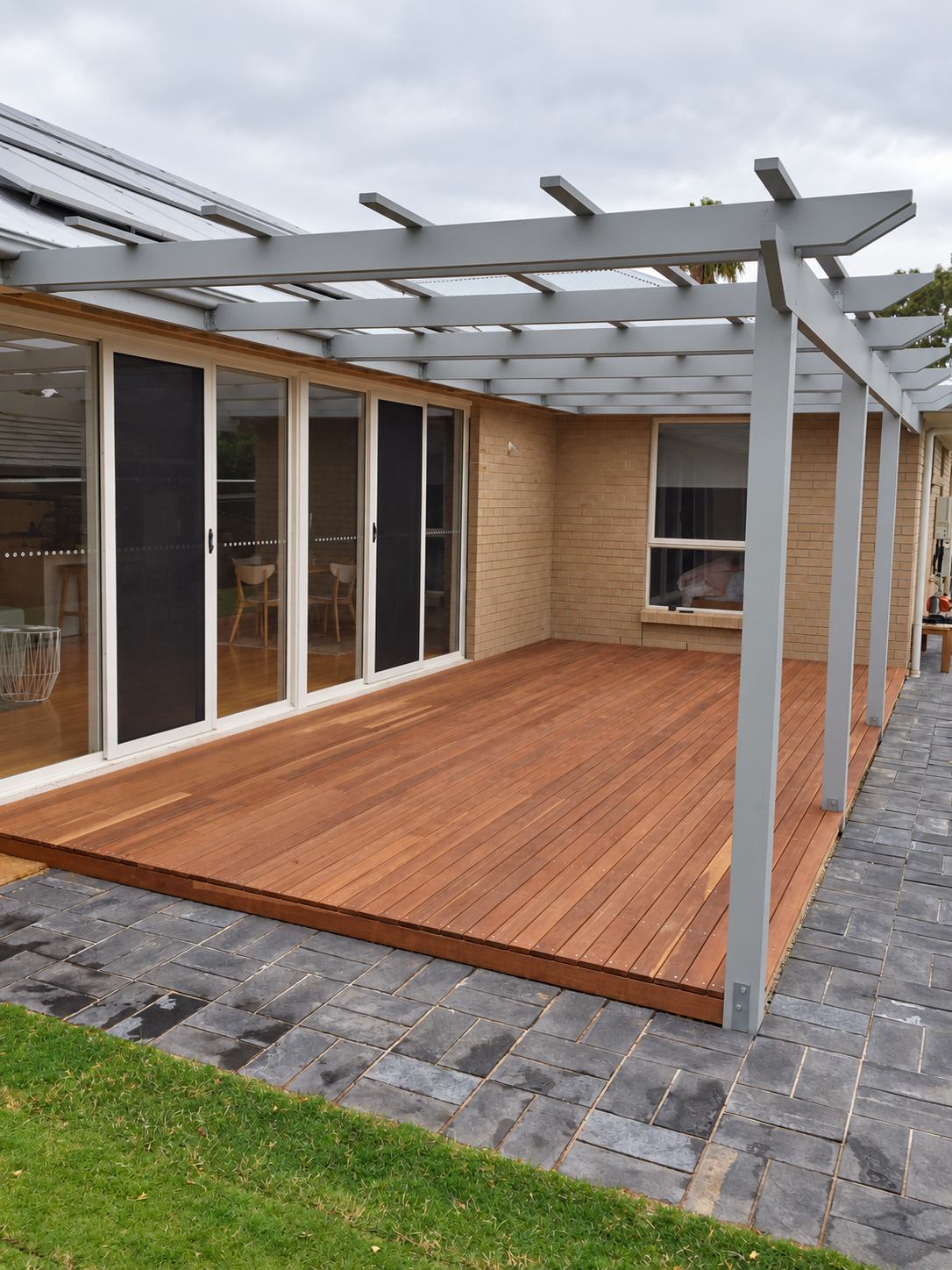 Modern backyard patio with a timber deck, gray pergola, sliding glass doors, brick house, and slate paving