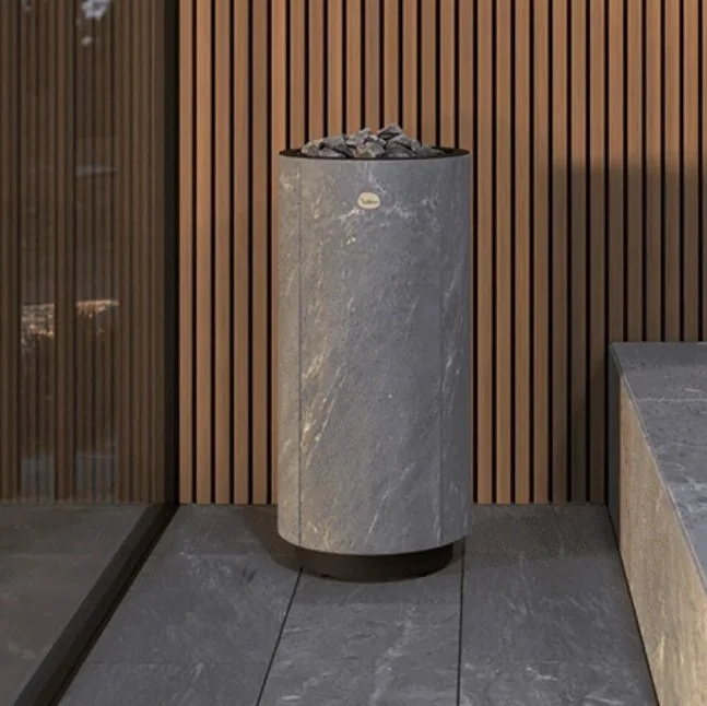 Cylindrical stone sauna heater filled with rocks against a vertical wood-slat wall