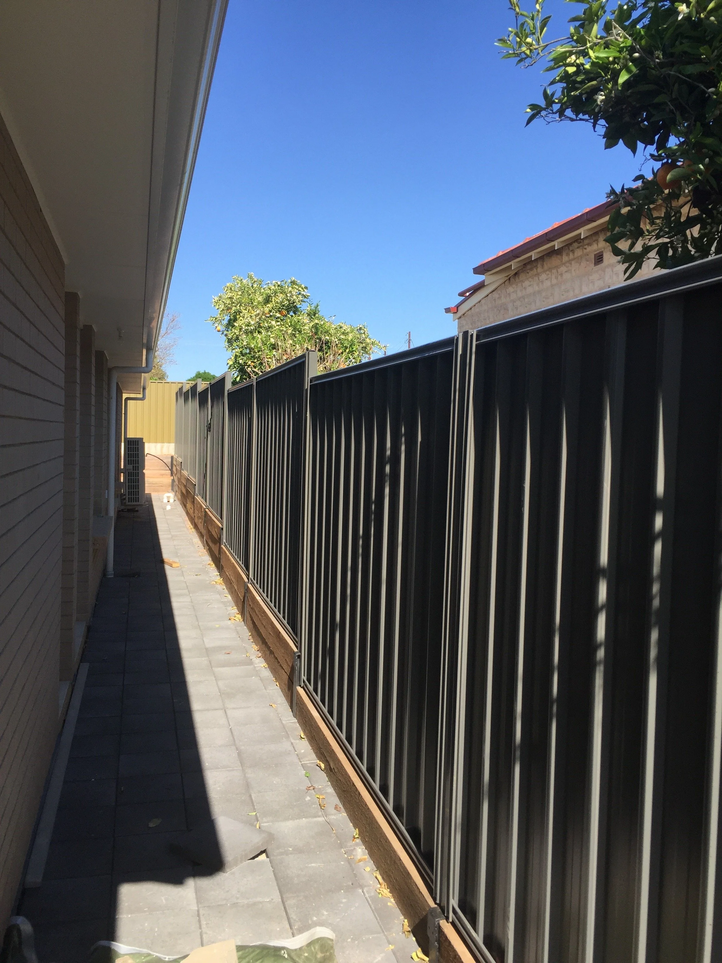 Narrow paved side yard between a house and black metal fence, with citrus trees, rooftops, and a clear blue sky.