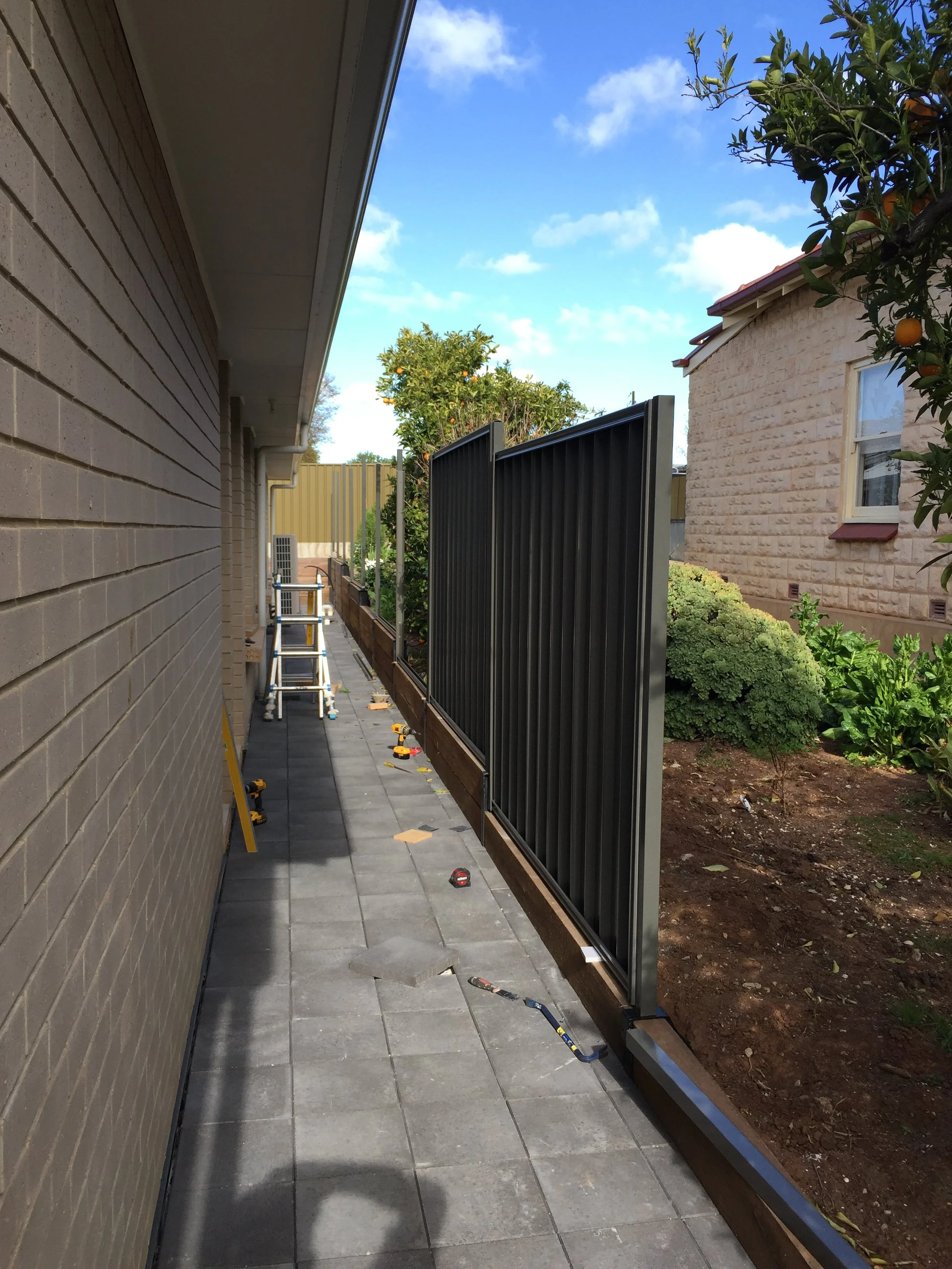 Side yard between brick homes with a black privacy fence, citrus trees, garden beds, patio pavers, and construction tools.