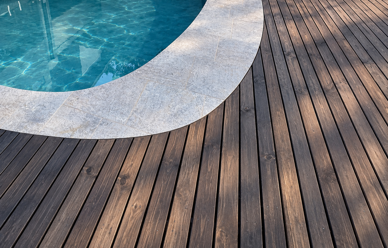 Curved swimming pool with turquoise water, stone tile edging, and dark wooden deck.