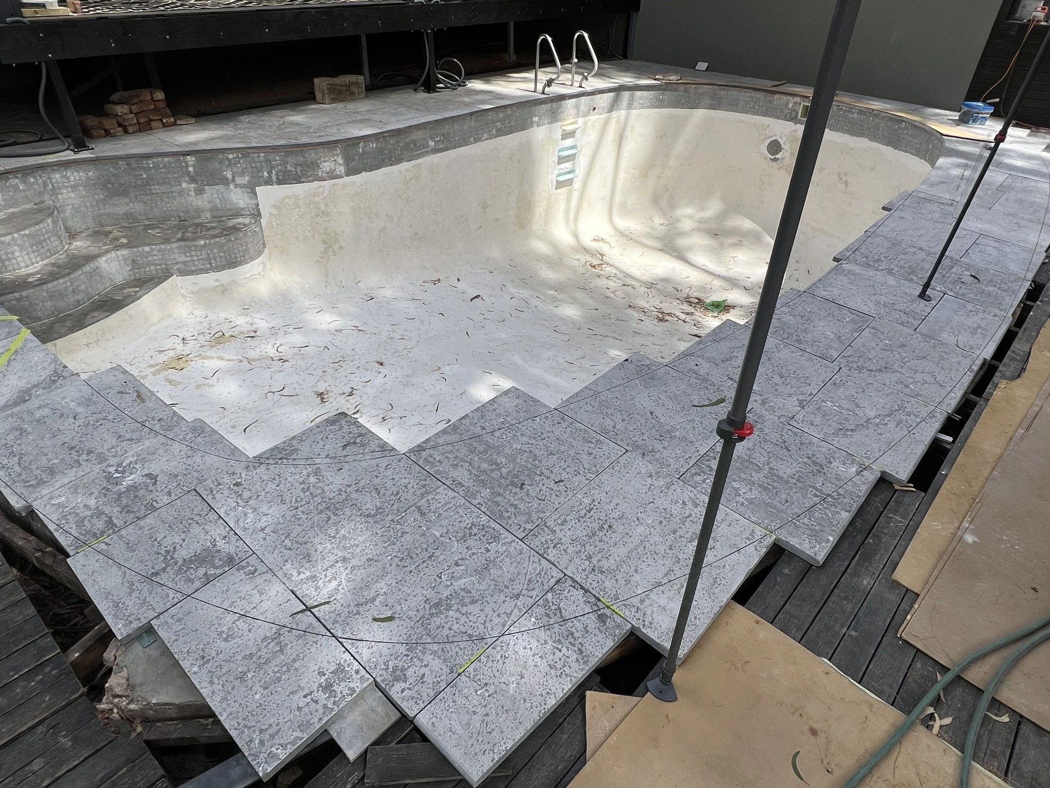 Empty in-ground swimming pool under renovation with exposed plaster, pool ladder, and gray patio tiles being installed around the deck.