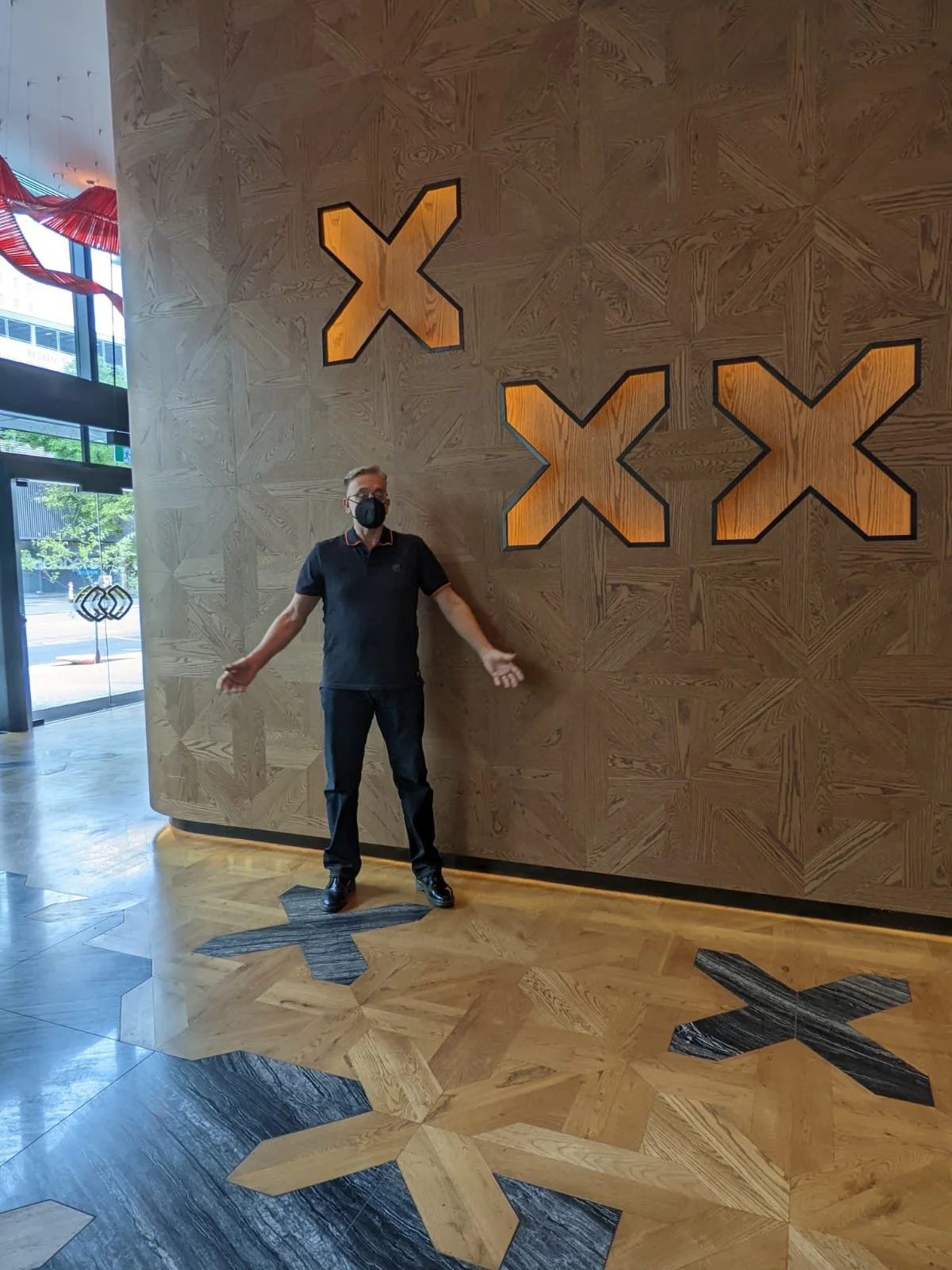 Masked man standing in a modern lobby beside illuminated wooden X-shaped wall artwork and matching patterned flooring