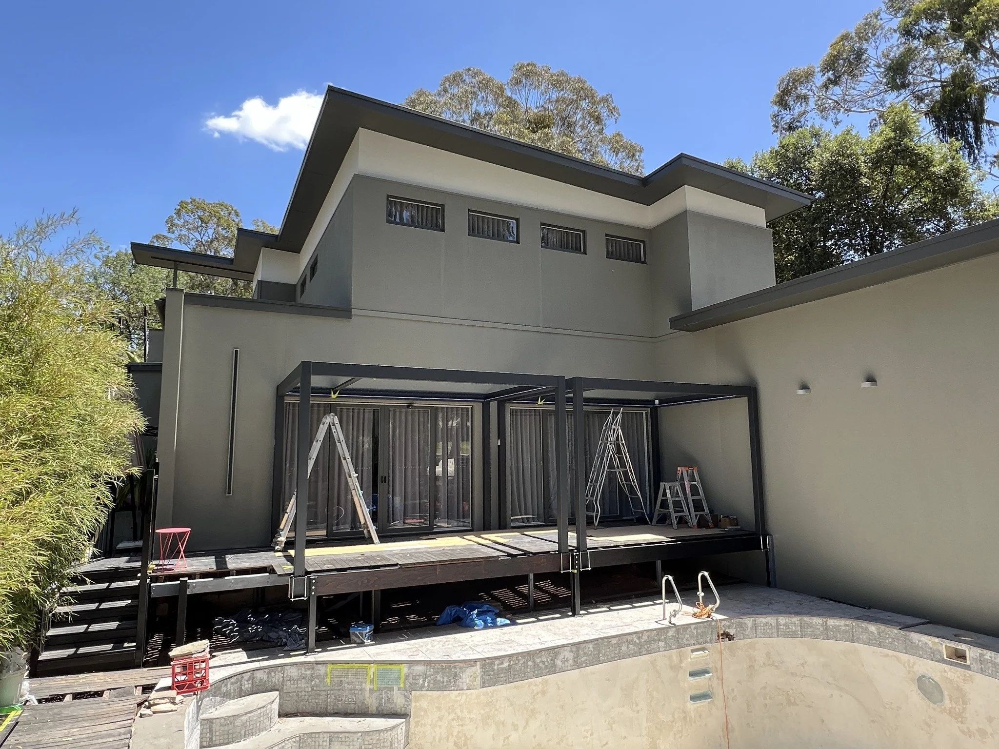 Modern two-story house with a covered patio, sliding glass doors, ladders, and an unfinished swimming pool in the backyard