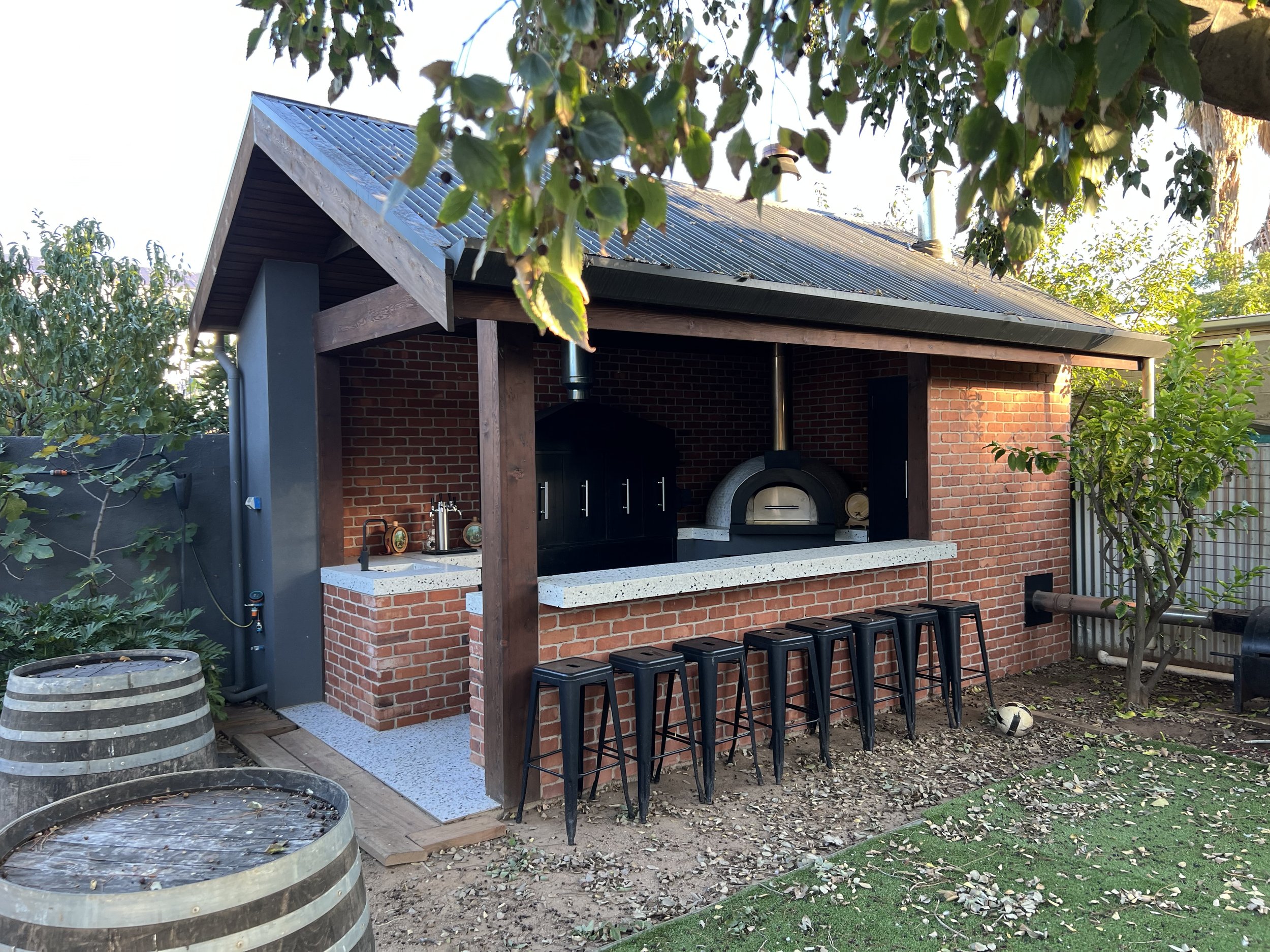 Outdoor brick entertainment area with a wood-fired pizza oven, bar counter, stools, and wine barrels beneath a covered patio