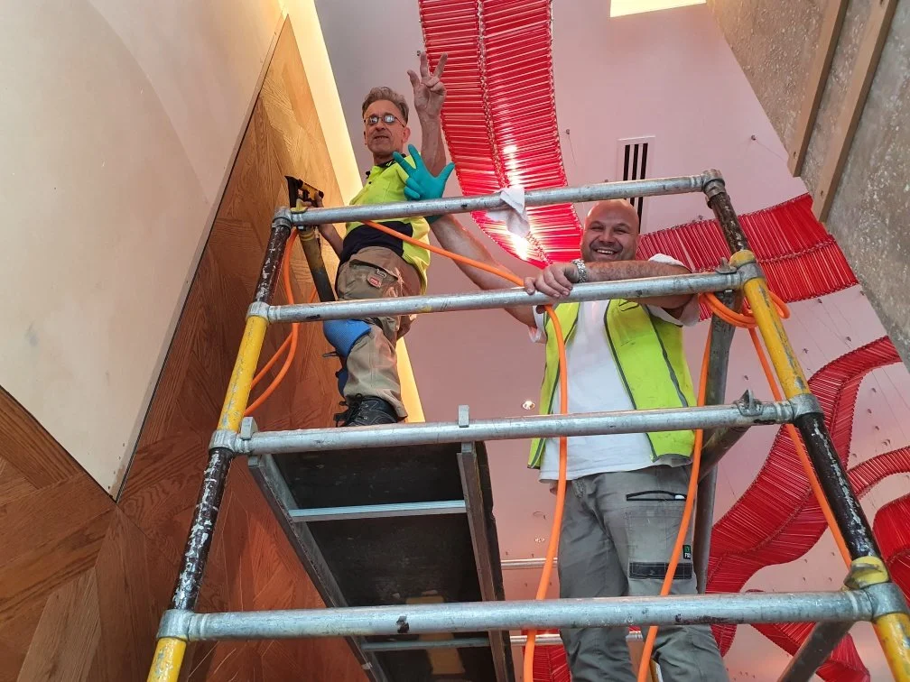 Two construction workers standing on a mobile scaffold inside a building, with red spiral ceiling decorations in the background.