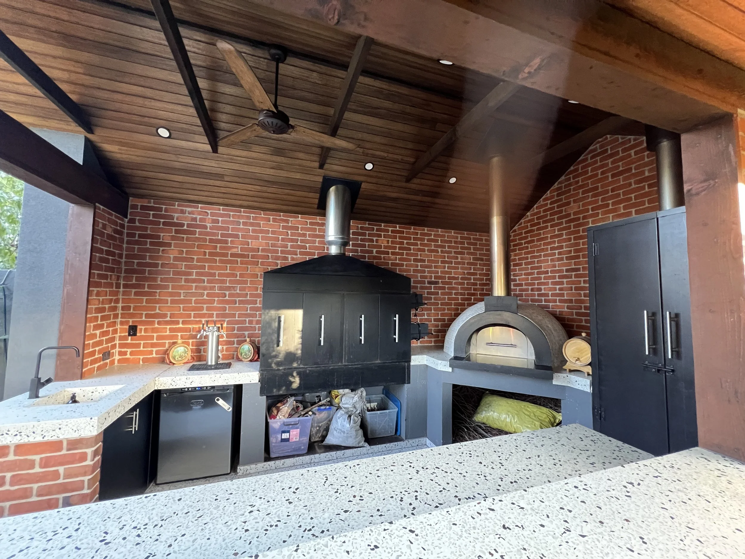 Outdoor brick kitchen with a wood-fired pizza oven, black grill, sink, refrigerator, wood ceiling, and ceiling fan
