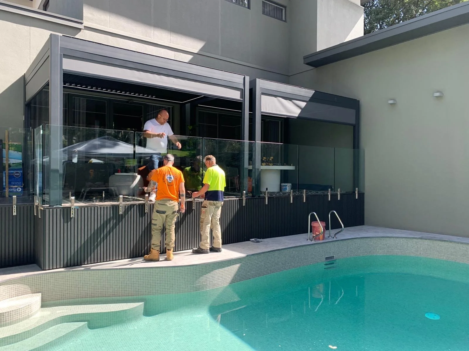 Three men inspect a glass pool fence beside a turquoise swimming pool at a modern home.
