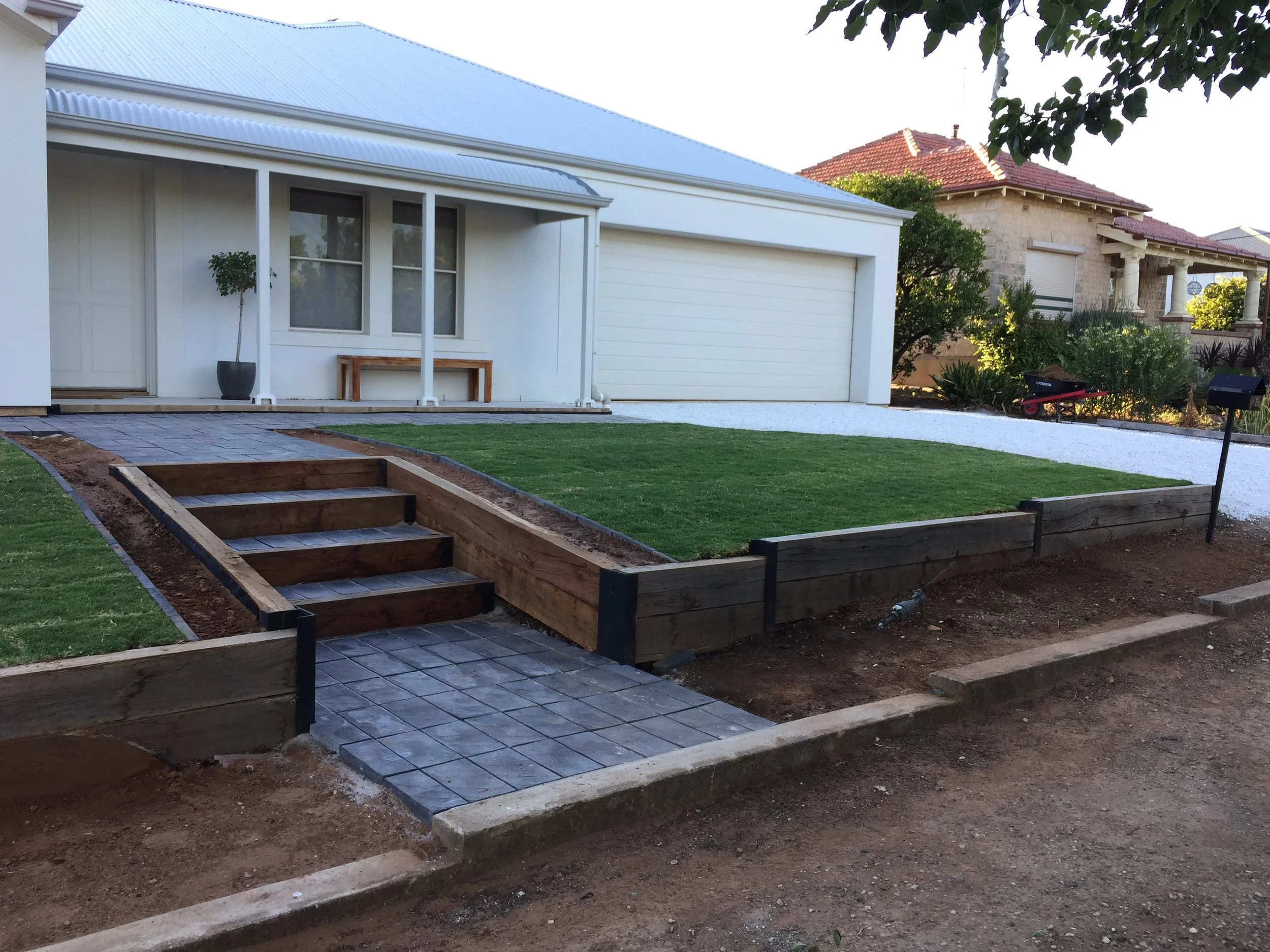 White single-story house with covered porch and garage, landscaped front yard, raised garden beds, paved walkway, and neighboring brick home