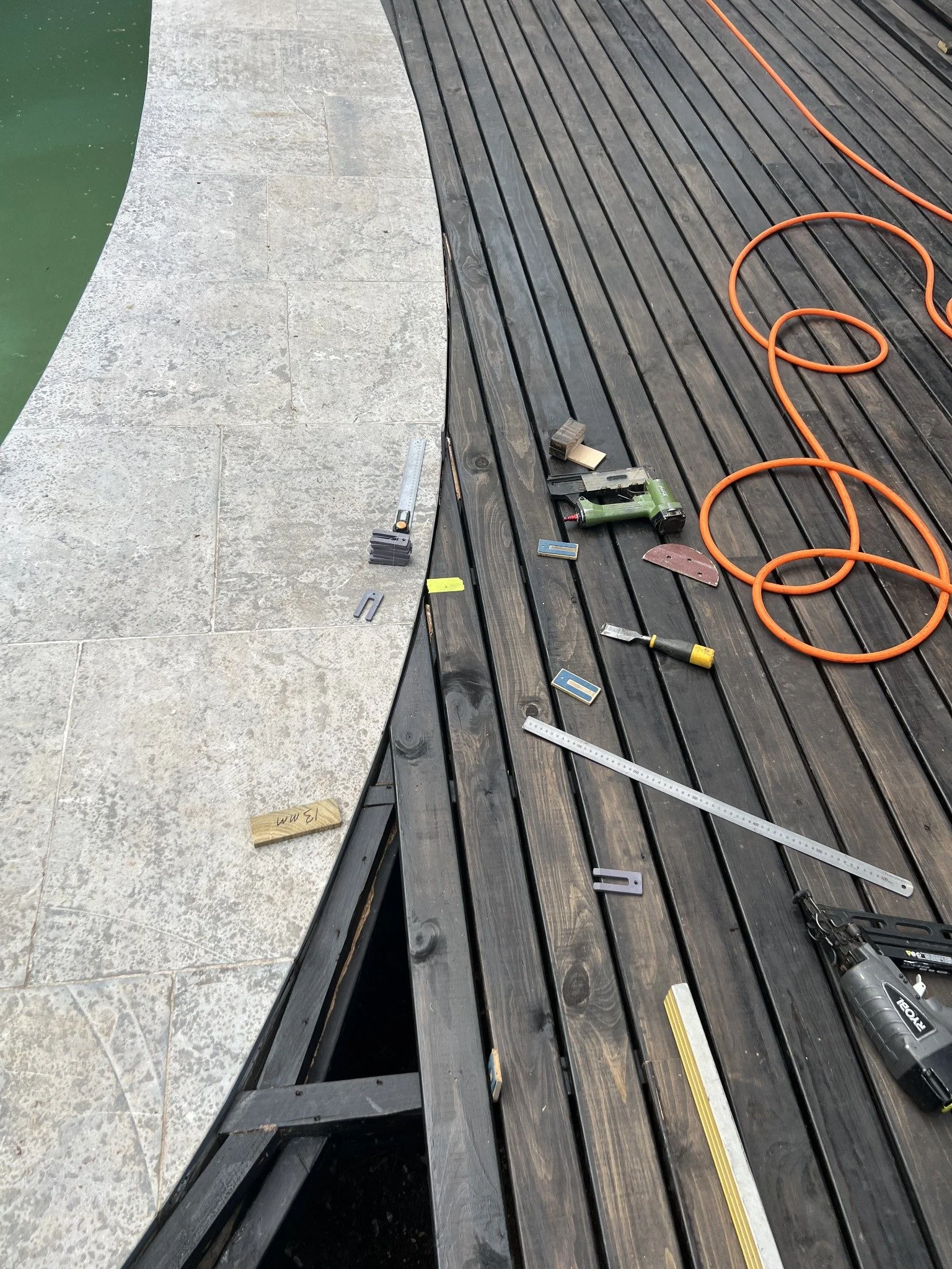 Tools and orange extension cord scattered across a dark wooden deck beside a stone-tiled pool edge