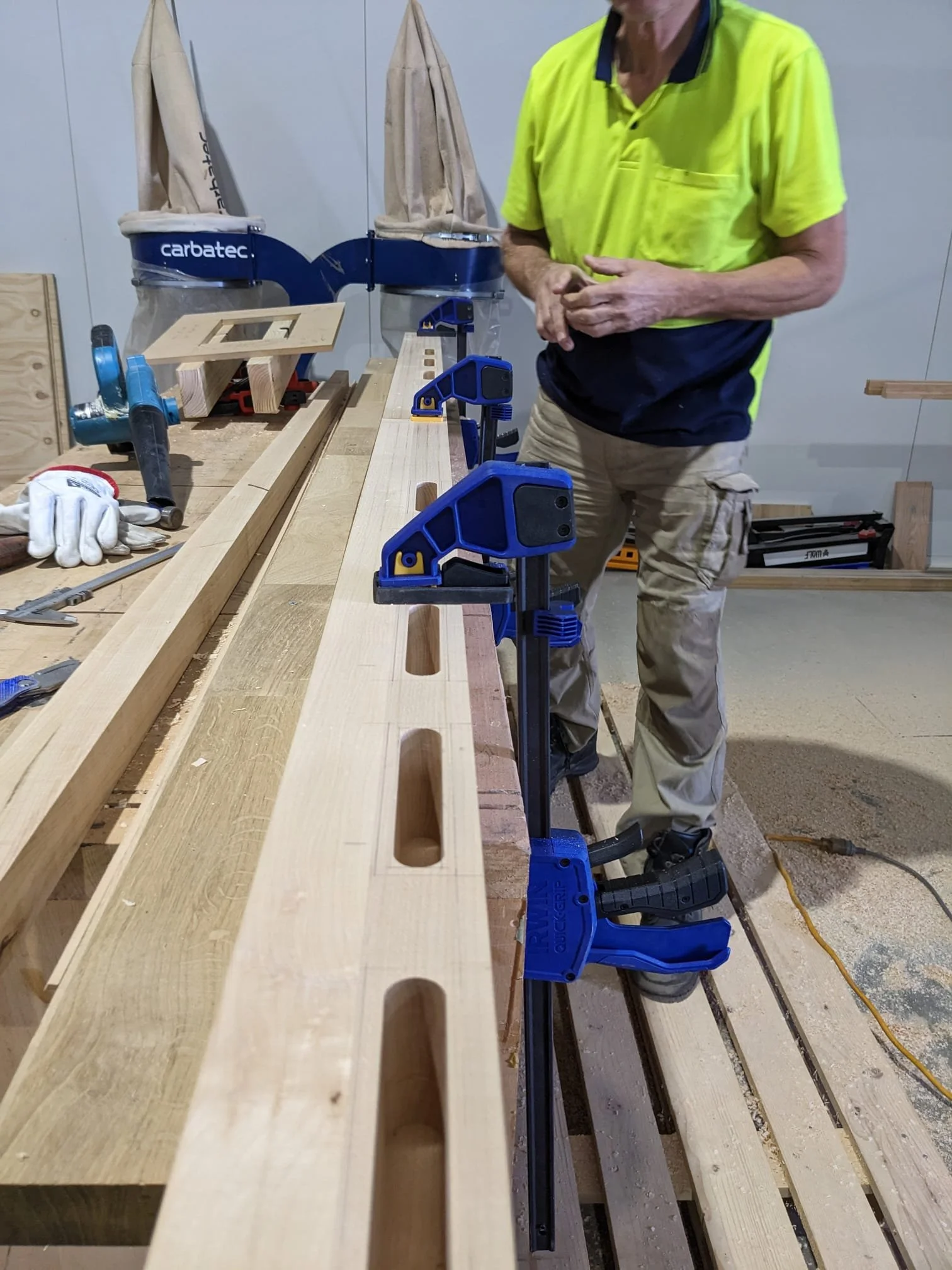 Woodworker in a workshop assembling a wooden project clamped to a workbench with blue clamps and power tools.