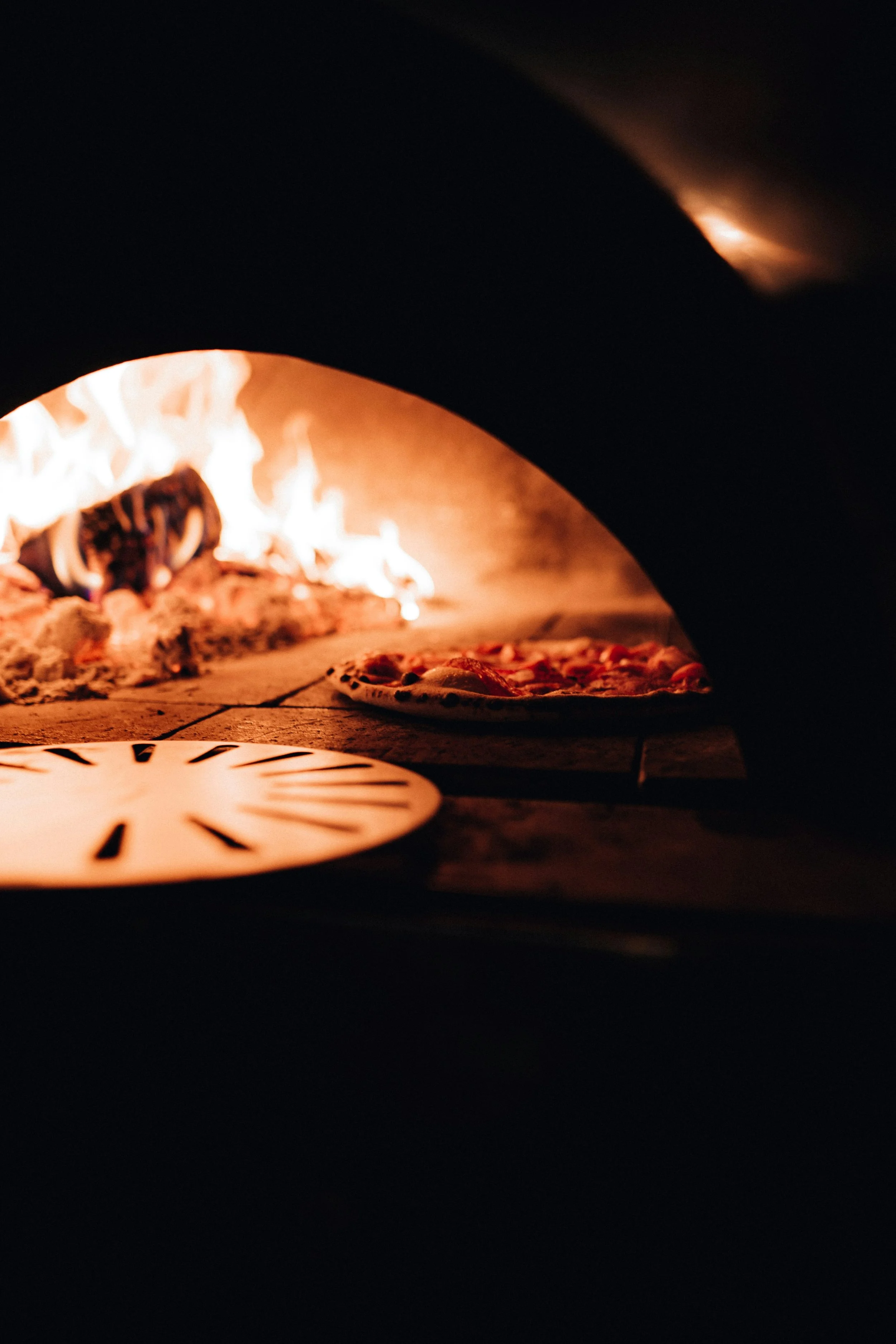 Close-up of a wood-fired pizza oven with flames, pizza, and a wooden pizza peel.