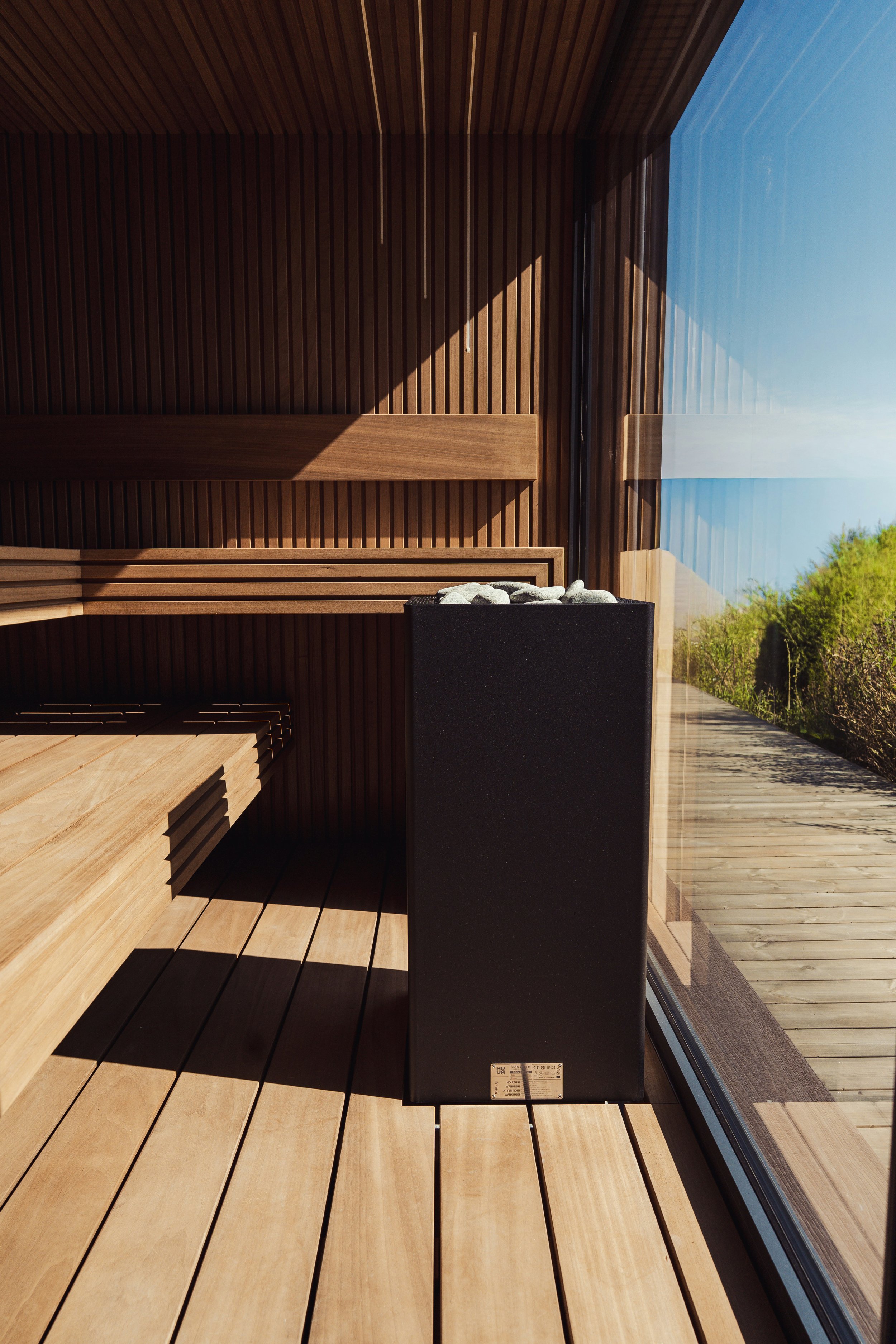 Modern wood-paneled sauna with benches, black stone-filled heater, glass wall, and outdoor deck